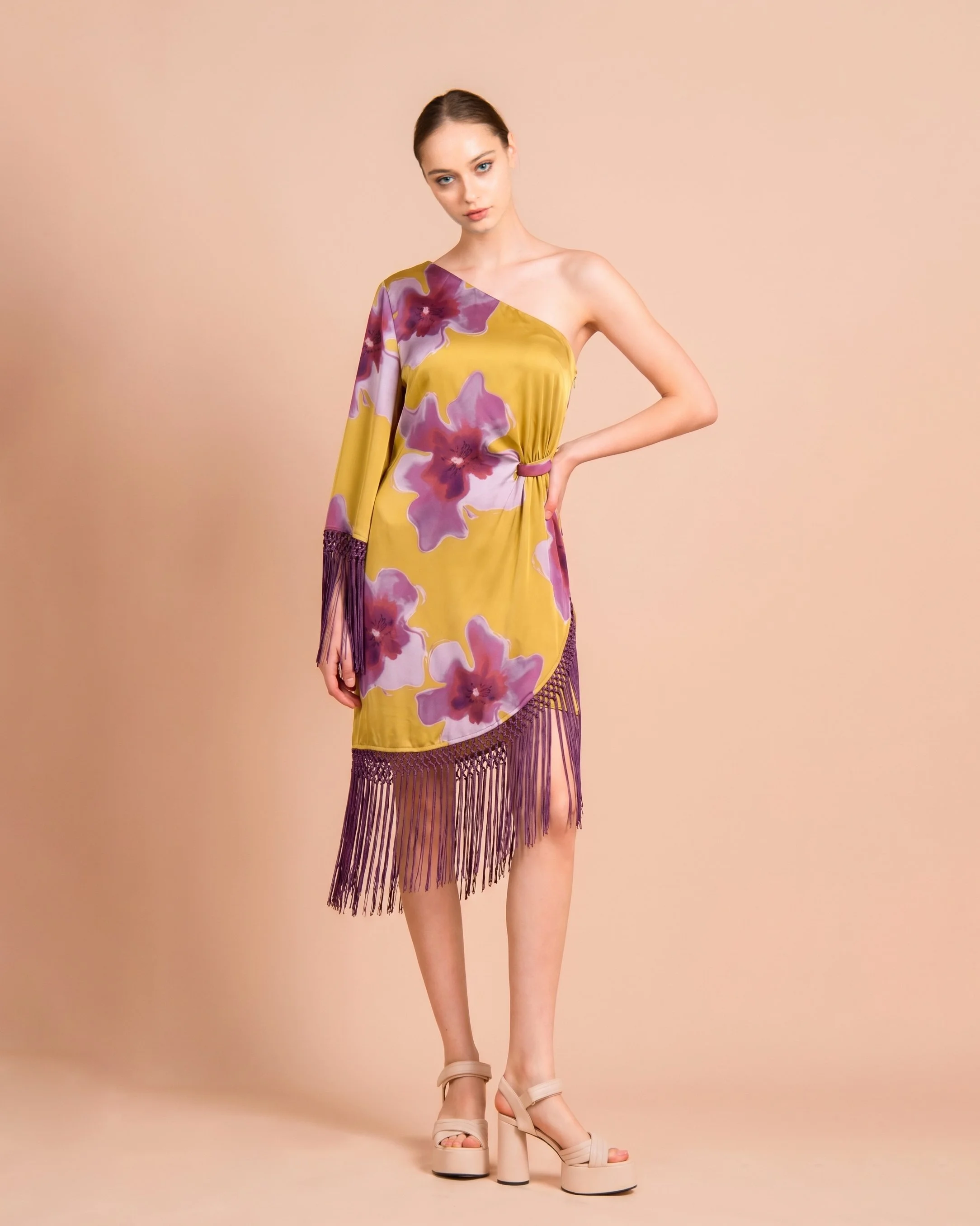 Young woman wearing an asymmetrical yellow dress with purple floral pattern, fringe detail at hem, and beige high-heeled sandals, posing against a plain beige background.