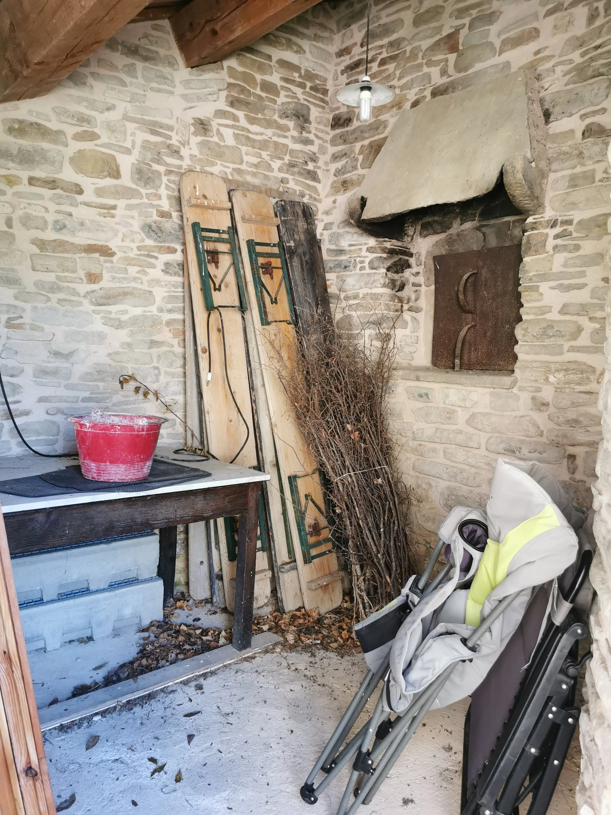 A rustic room with a stone wall, fireplace, and wooden ceiling beams. Various sleds and wooden planks are leaning against the wall. There is a table with a red bucket and some tools, a pile of leafless branches, a stroller, and leaves on the ground.