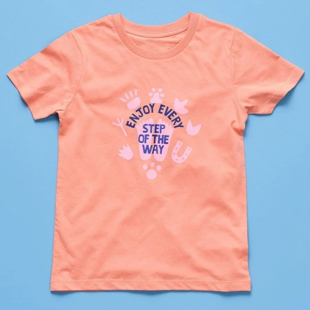 Peach-colored T-shirt with a printed message and graphics in pink and dark blue, centered on a light blue background.