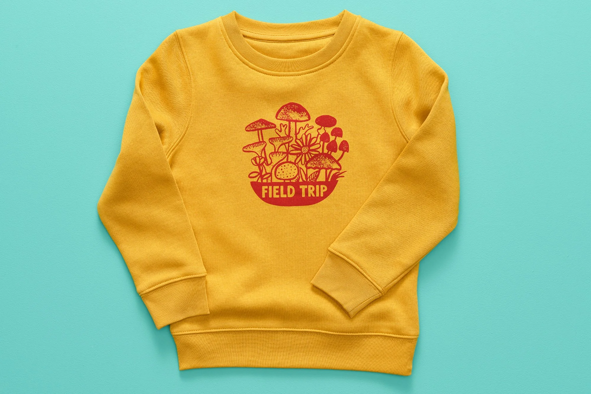Yellow sweatshirt with a red graphic of mushrooms, flowers, and the words 'Field Trip' on the front, laid flat against a turquoise background.