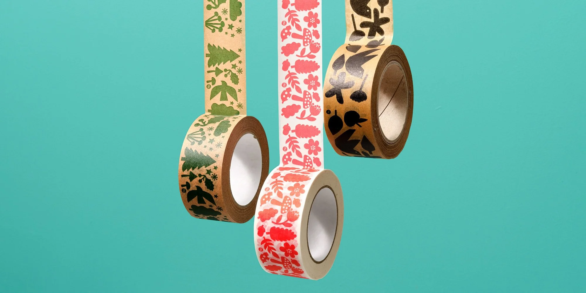 Four rolls of decorative washi tape featuring festive patterns, including Christmas trees, gingerbread cookies, and snowflakes, suspended against a teal background.