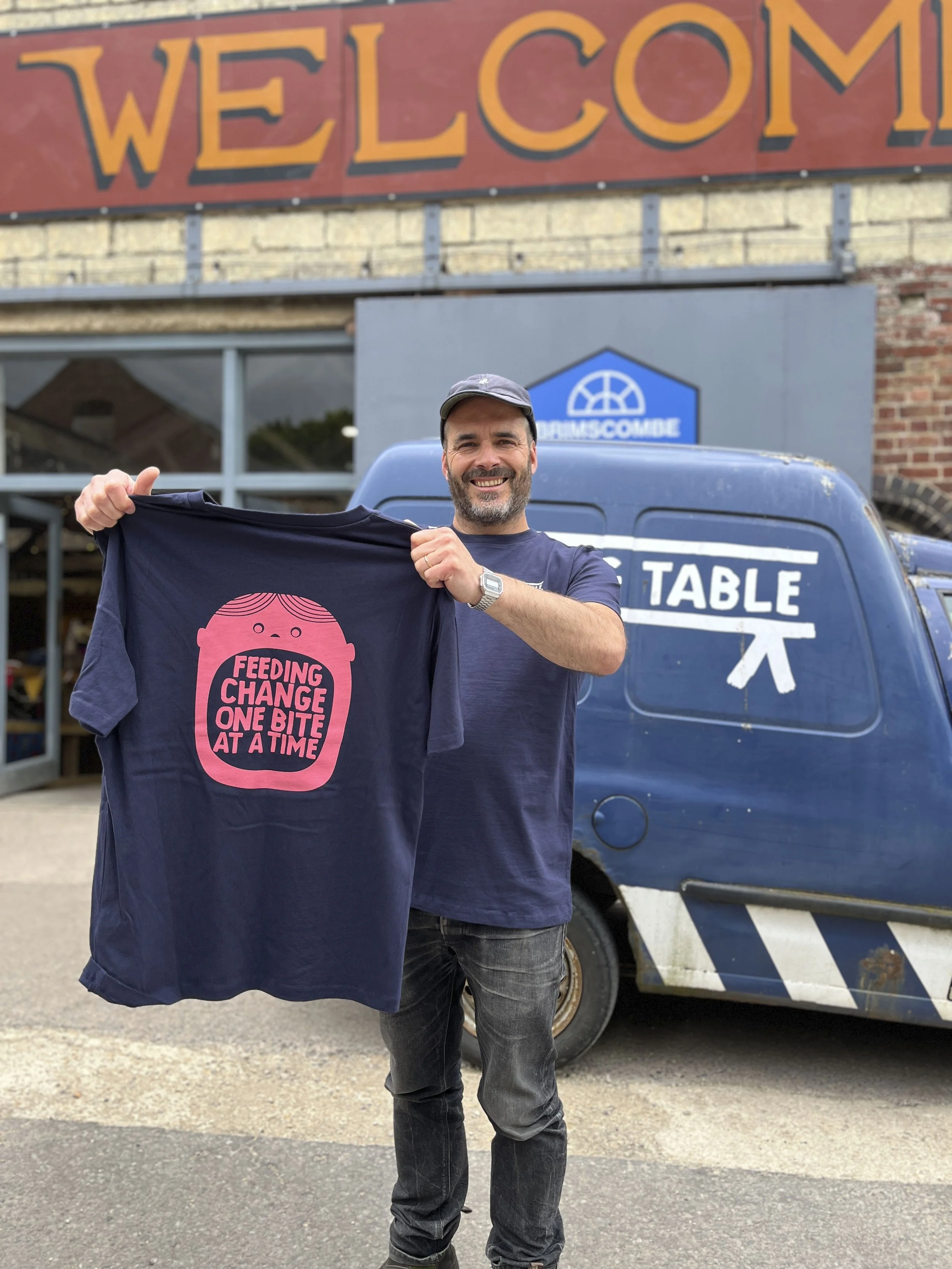 Man smiling and holding a dark blue t-shirt with a pink graphic of a baby's face and the text "FEEDING CHANGE ONE BITE AT A TIME". He is standing outside near a blue van with white graphics and text, with a brick building and large welcome sign in the background.