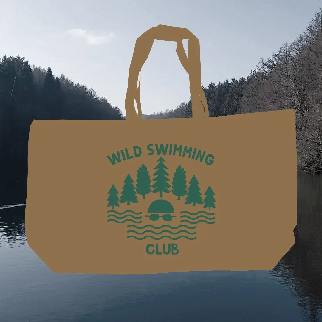 A brown paper shopping bag with a logo that reads "Wild Swimming Club" and features trees, water, a turtle wearing glasses, and an umbrella, set against a background of a river and trees on a cloudy day.