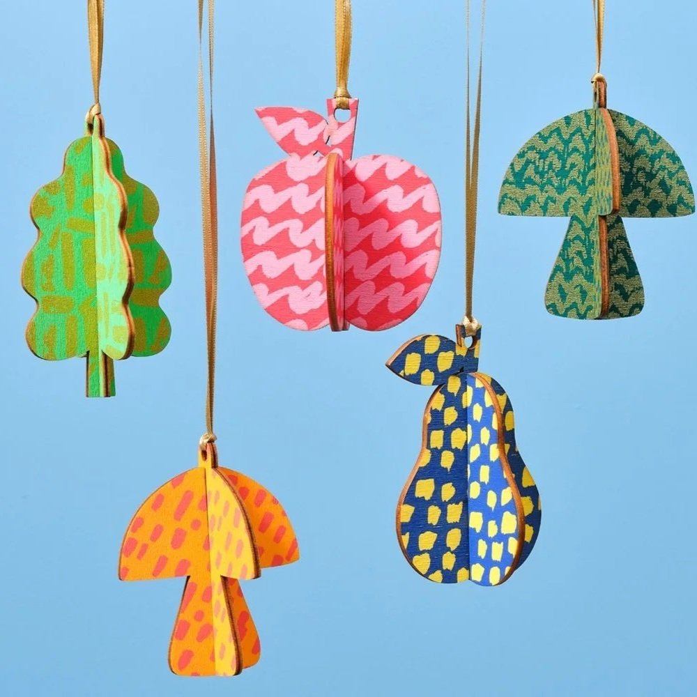 Colorful paper zippered ornaments shaped like fruits and vegetables, including a green leafy vegetable, pink peach, green mushroom, yellow pear, and blue and yellow ladybug, hanging against a blue background.