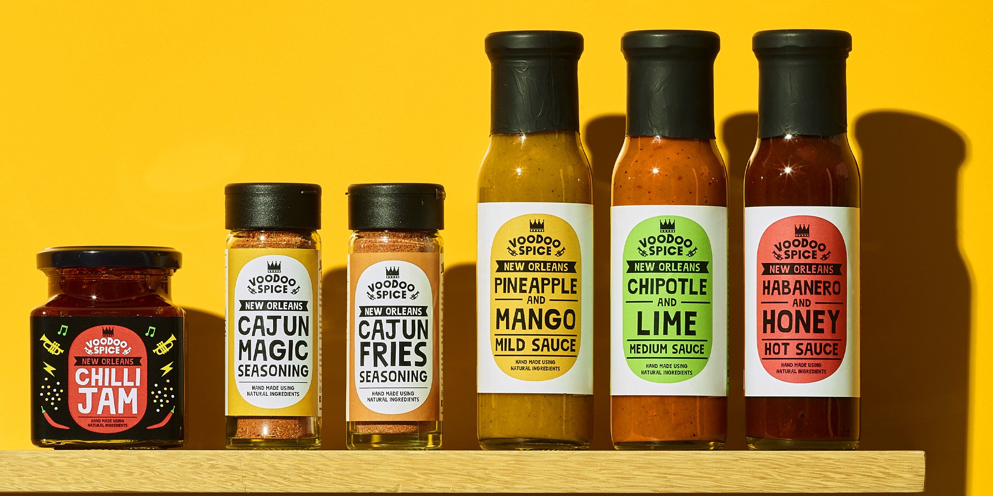 Six bottles and jars of Voodoo Spice seasoning and sauces on a wooden surface against a yellow background. The products include Chili Jam, Cajun Magic Seasoning, Cajun Fries Seasoning, Pineapple and Mango mild sauce, Chipotle and Lime medium sauce, and Habanero and Honey hot sauce.