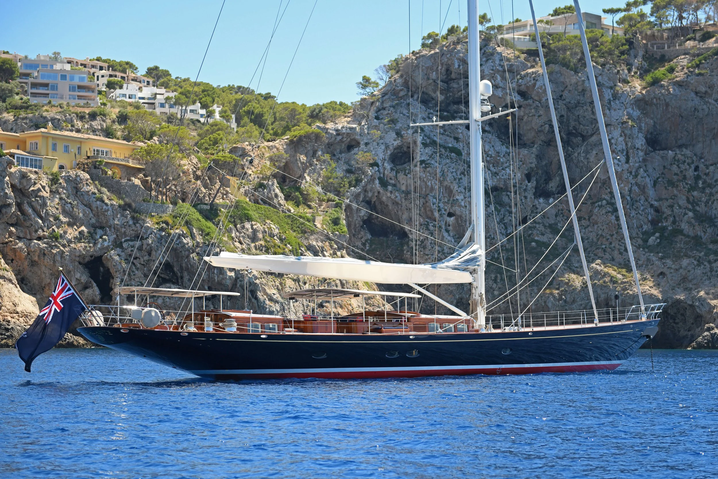 A sailboat with a dark blue hull and white sails floating on calm water near rocky coastline with hillside houses and greenery.