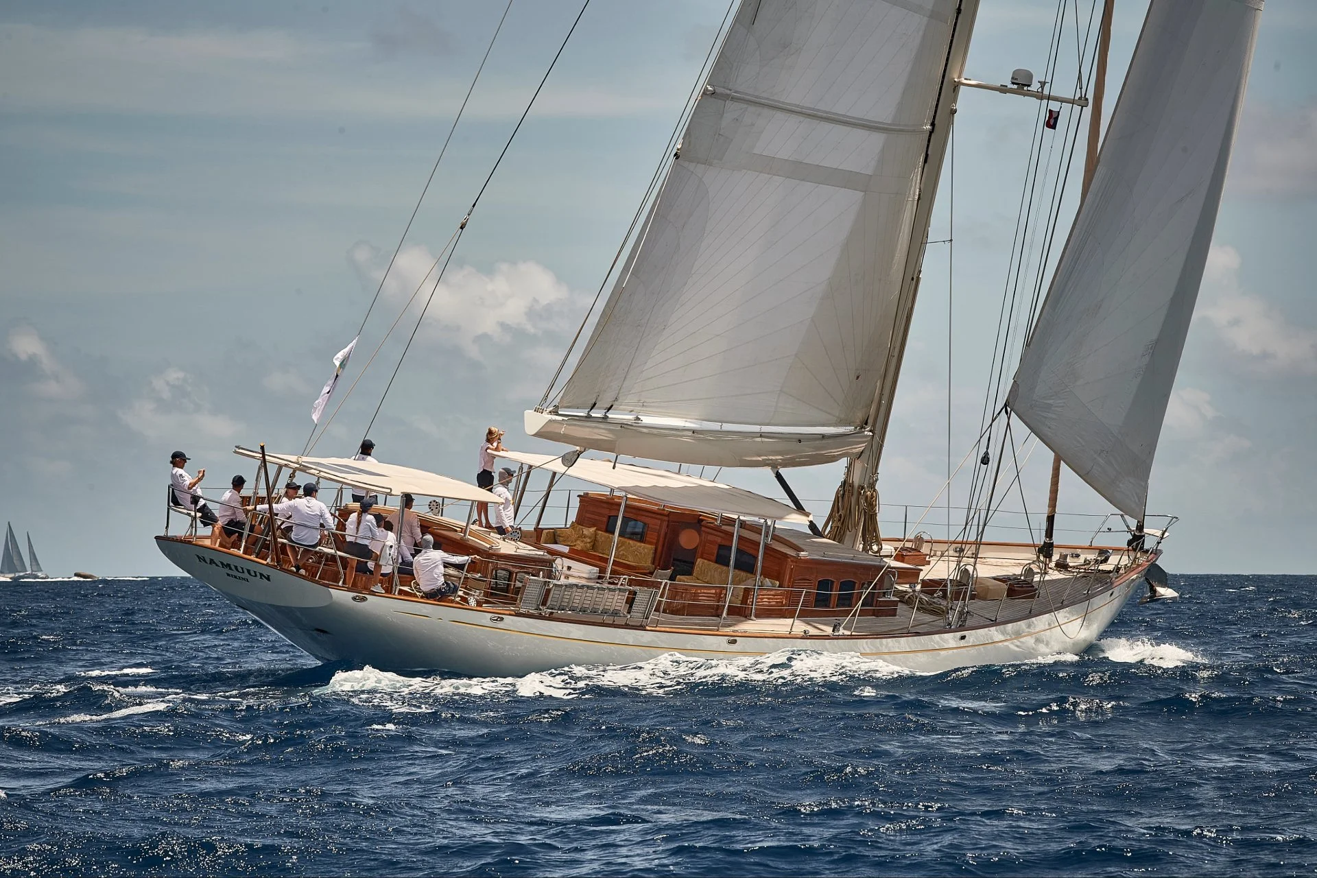 A sailing yacht named Namun with multiple crew members on board, some wearing white shirts, sailing on the ocean with a partly cloudy sky in the background.