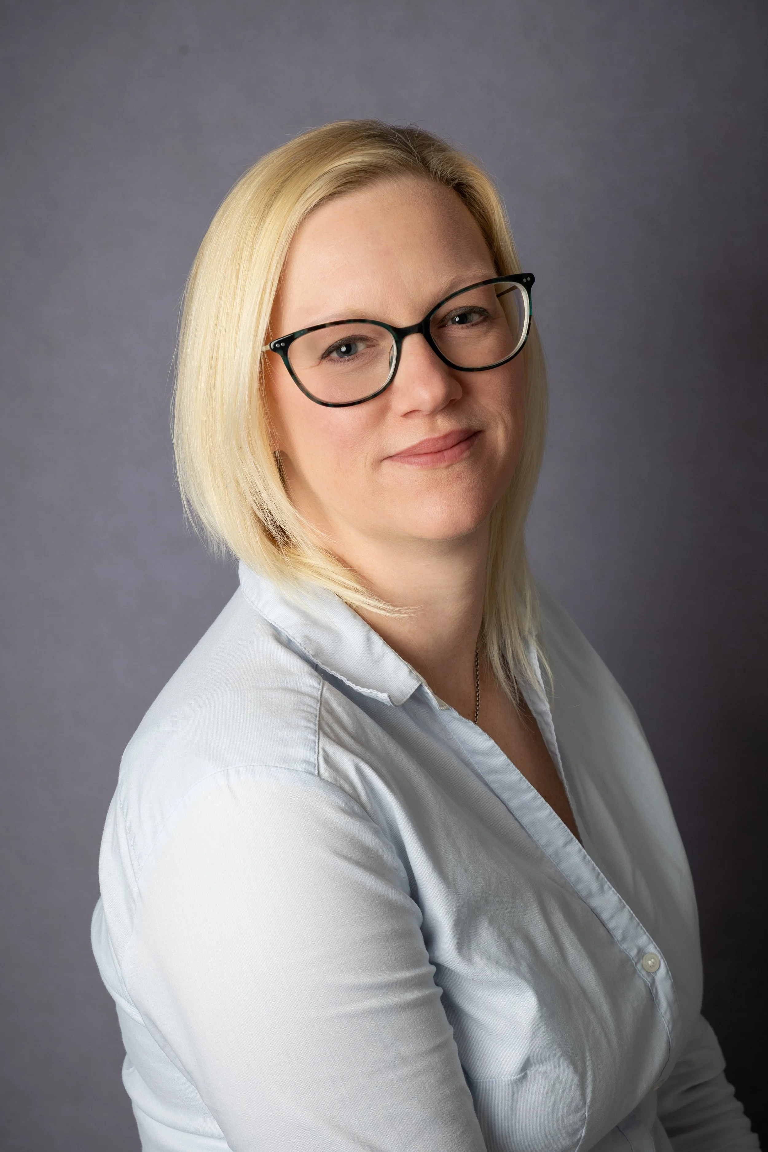 A woman with blonde hair, wearing glasses and a white collared shirt, smiling slightly against a gray background.