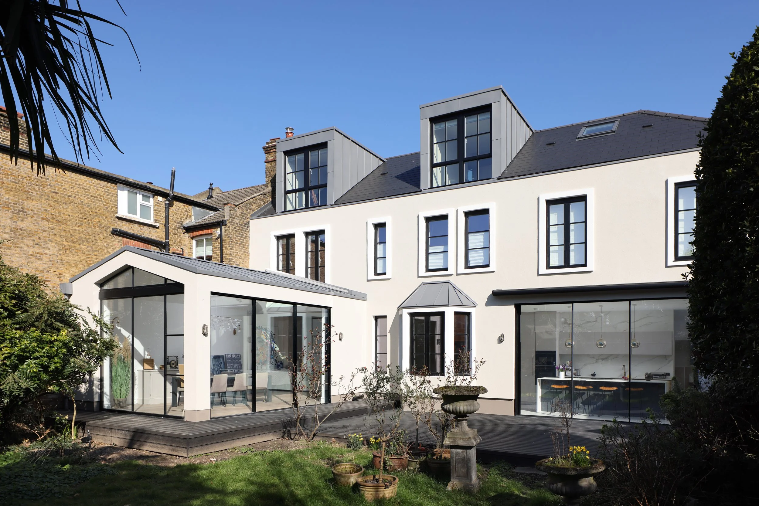Modern white house with large glass windows and a upper-floor dormer windows, situated in a garden with small trees and potted plants, under a clear blue sky.