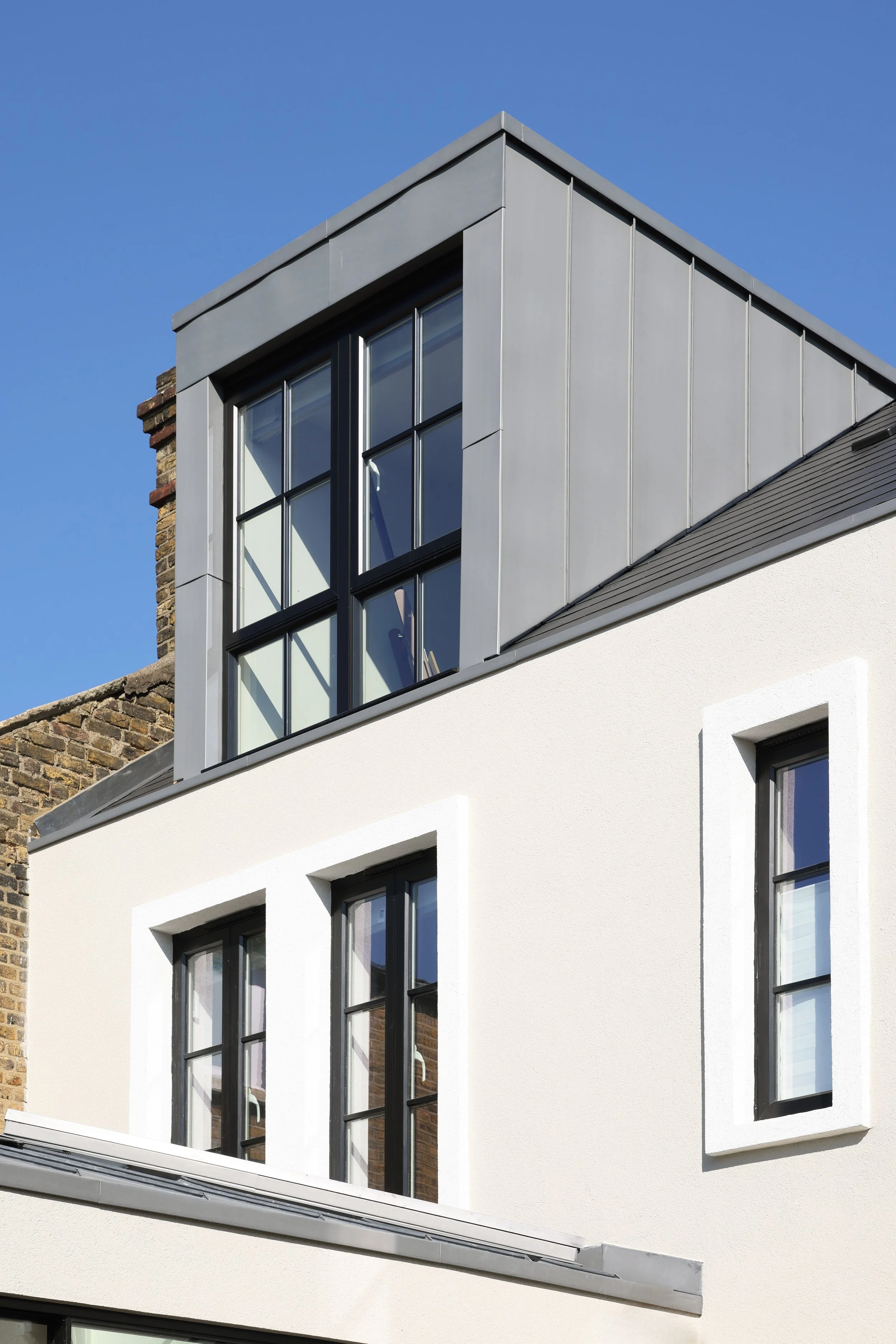 Modern building with large black-framed windows, white stucco exterior, and a gray metal roof section against a clear blue sky.