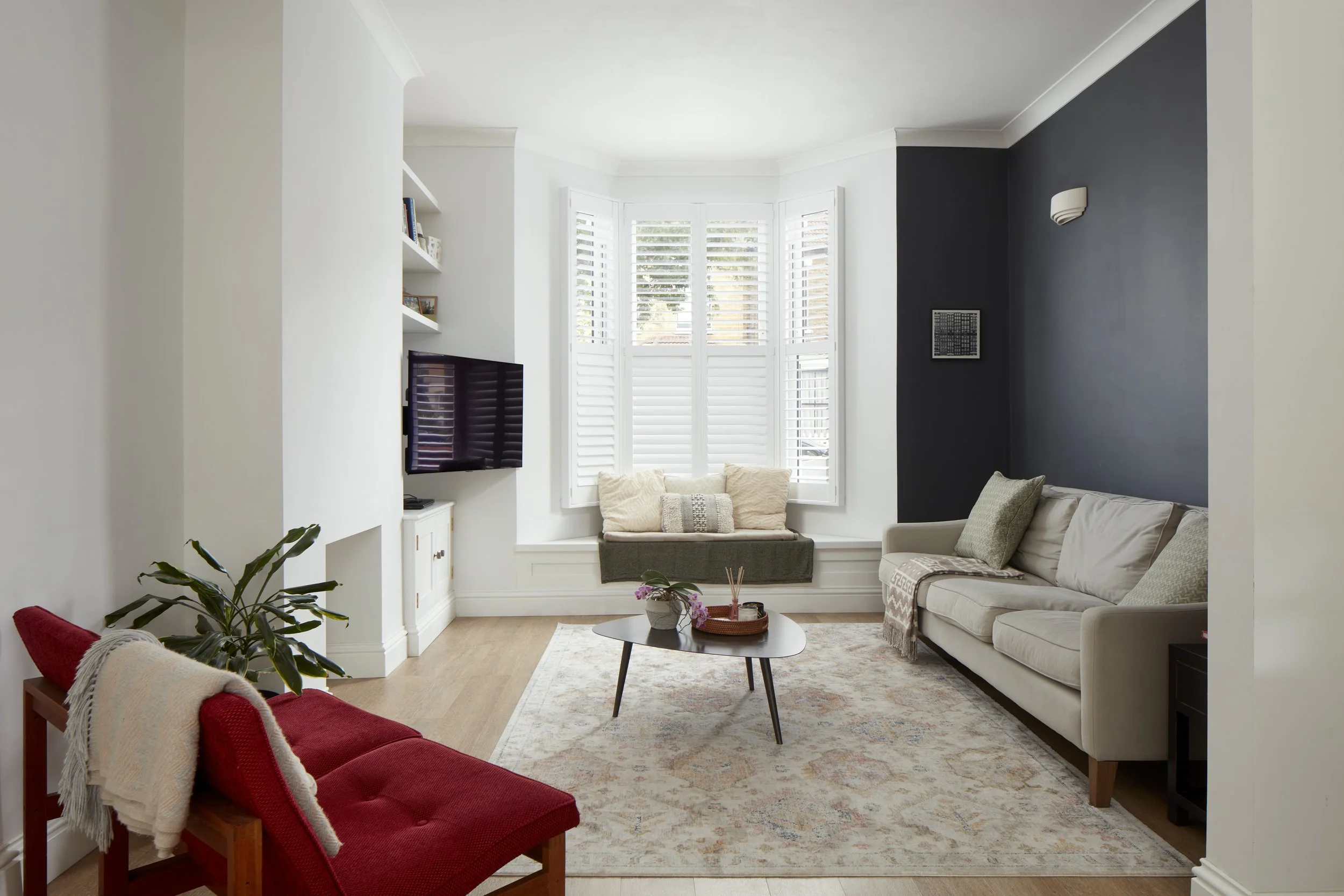 Living room with a beige sofa, a red armchair with a blanket, a coffee table with decorative items, a window with shutters, a wall-mounted TV, and a dark accent wall.