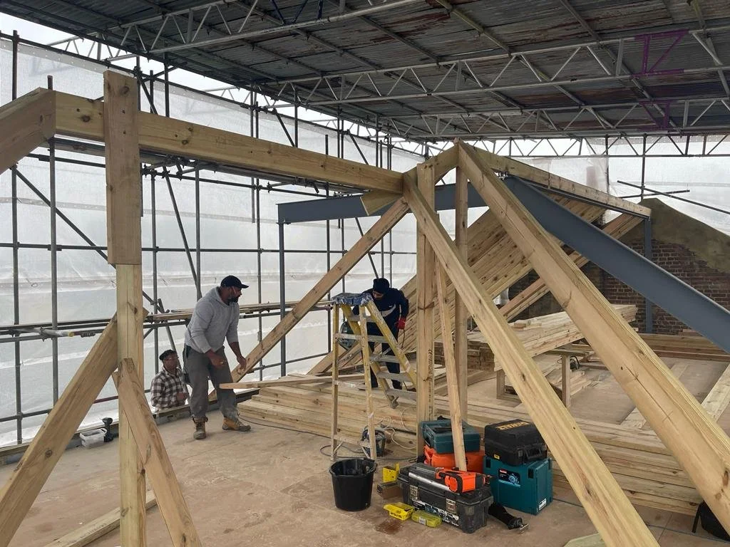 Construction workers building the wooden roof framework inside a partially enclosed structure with scaffolding and tools around.