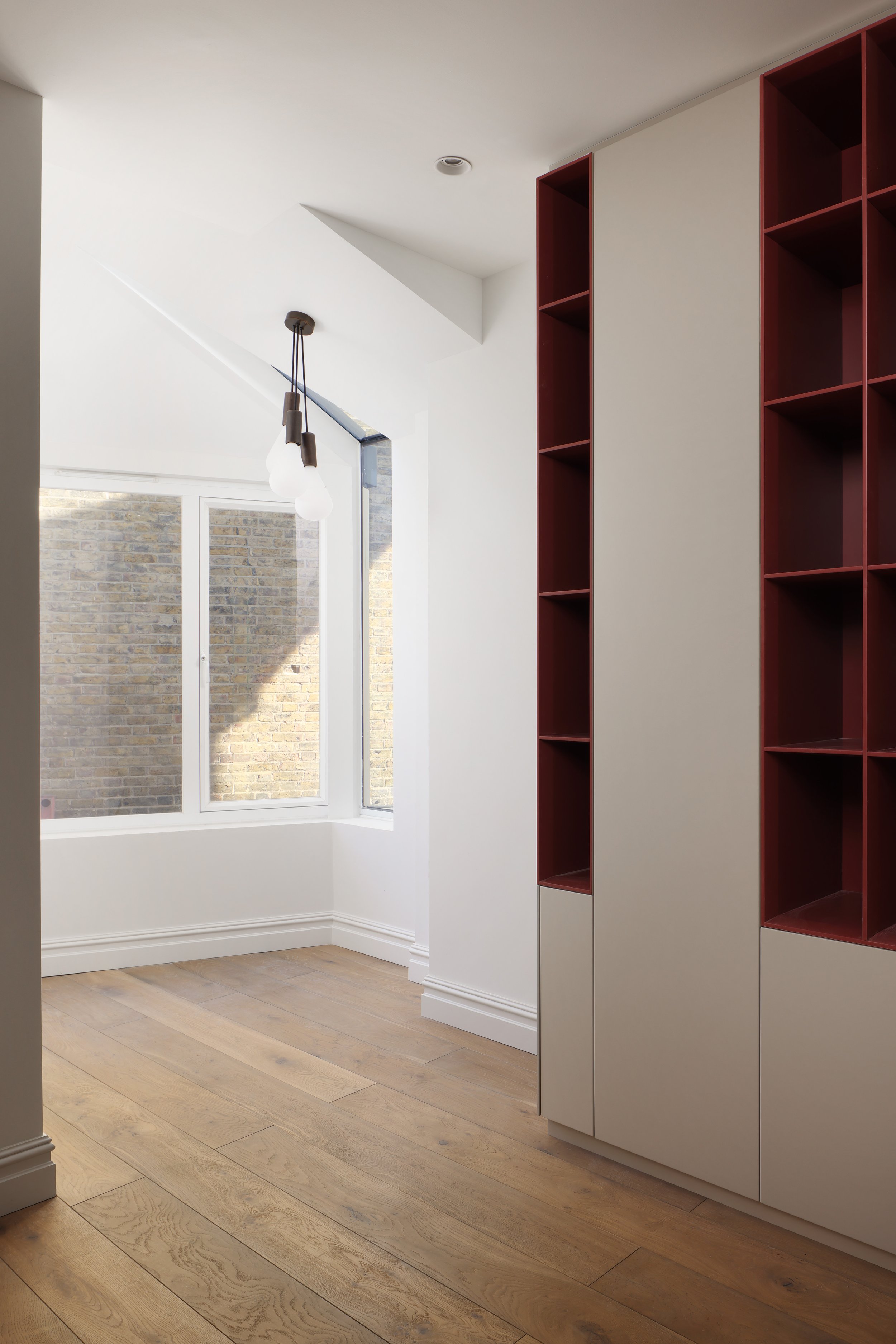 Empty room with wooden flooring, white walls, large window, modern hanging light fixture, red and beige built-in shelving unit.