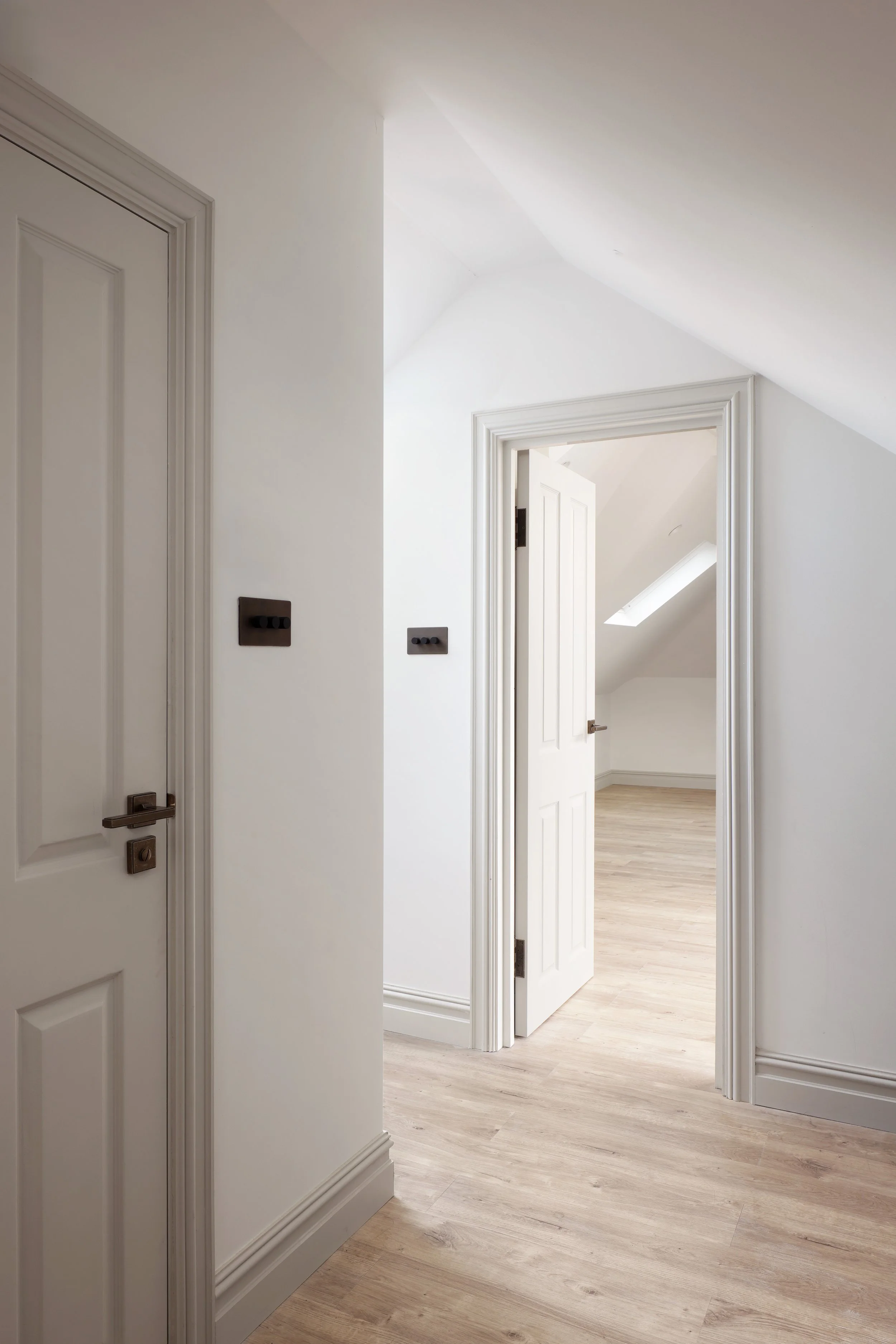 Image of a hallway with white walls, white doors, and light wood flooring. One door is open, revealing a room with a skylight.