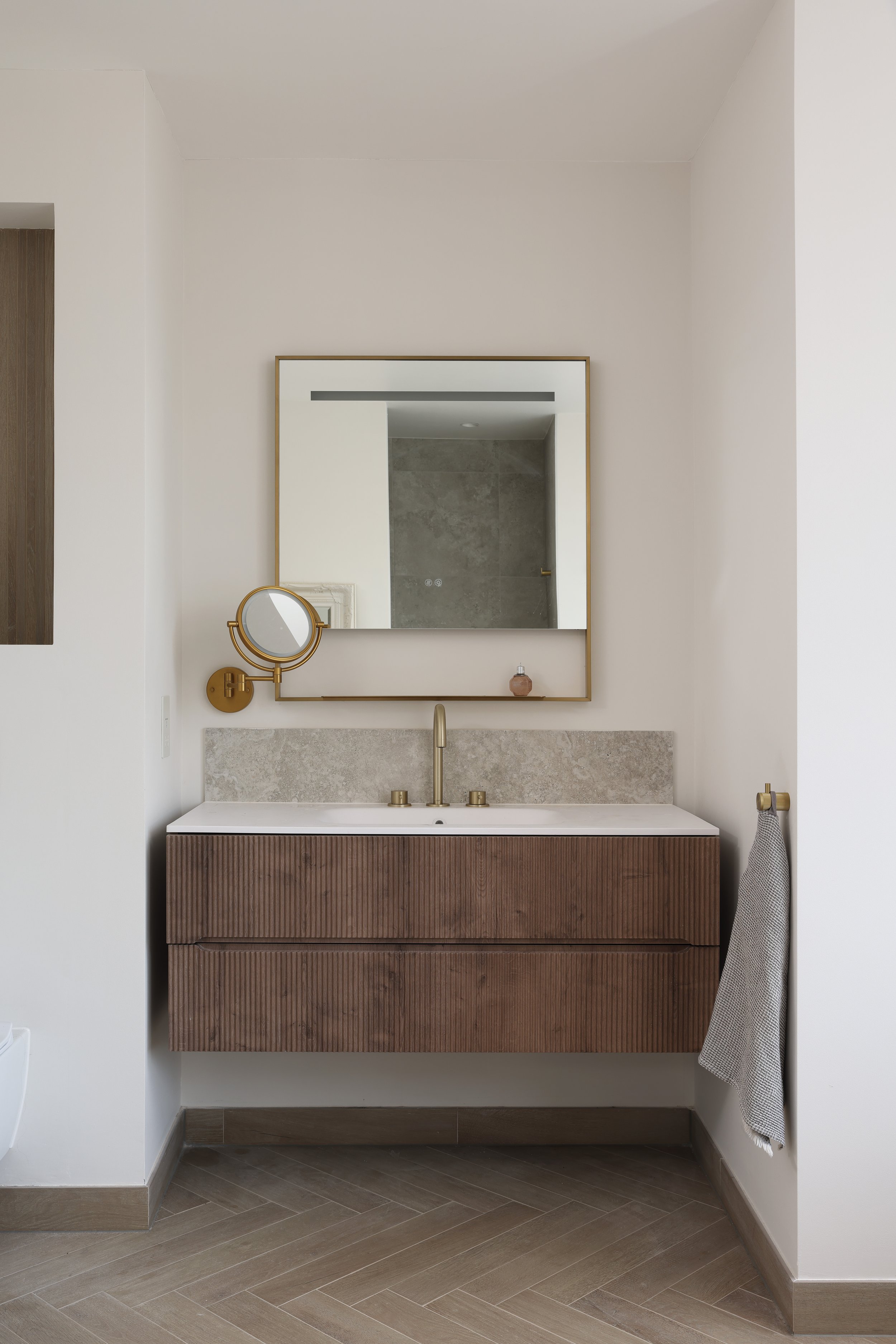 Modern bathroom vanity with a rectangular mirror, round magnifying mirror, and a beige countertop with a brass faucet, set against white walls with gray floor tiles.