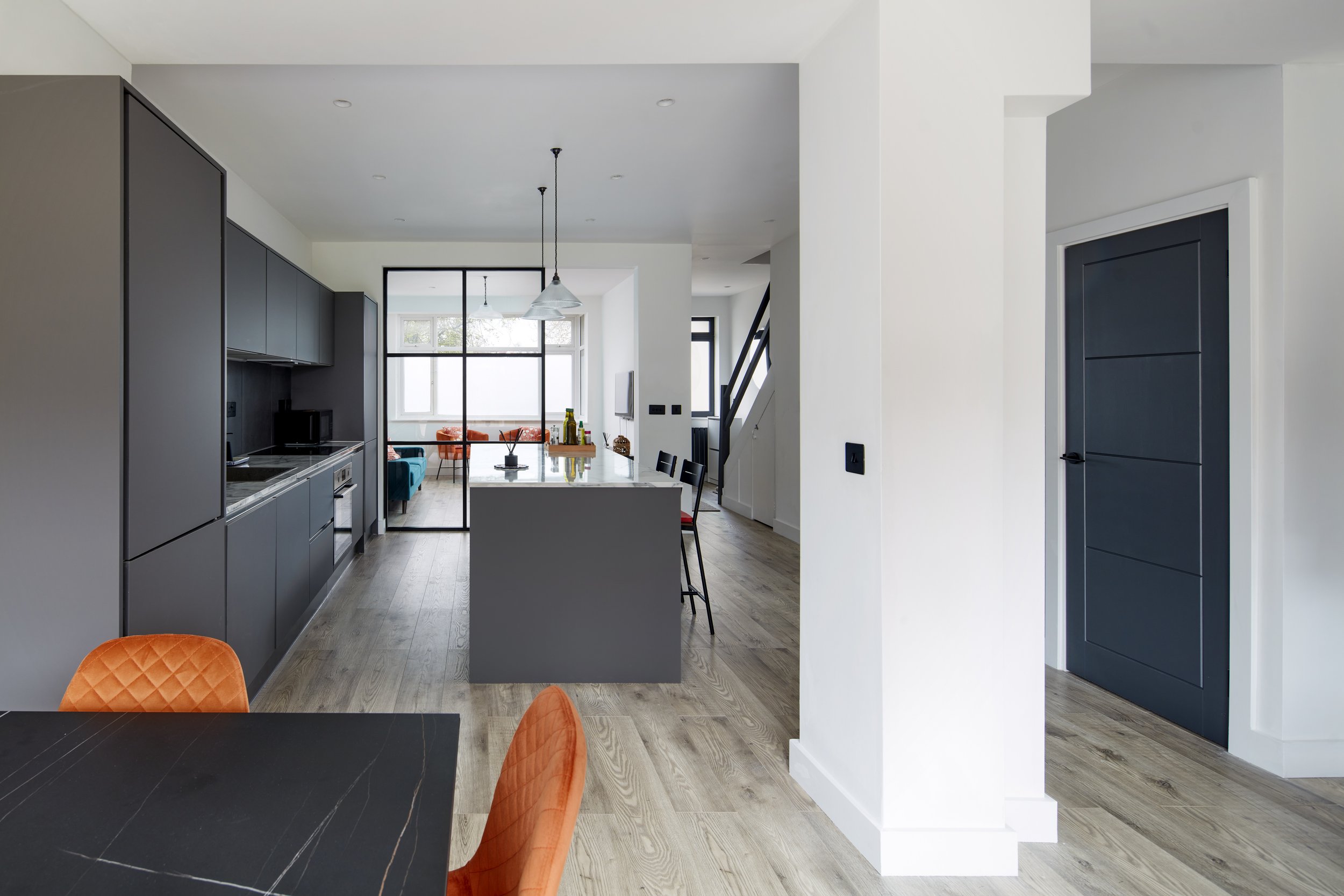 Modern open-plan kitchen and dining area with black cabinetry, a kitchen island, orange quilted chairs, and a sunlit living space with large windows.