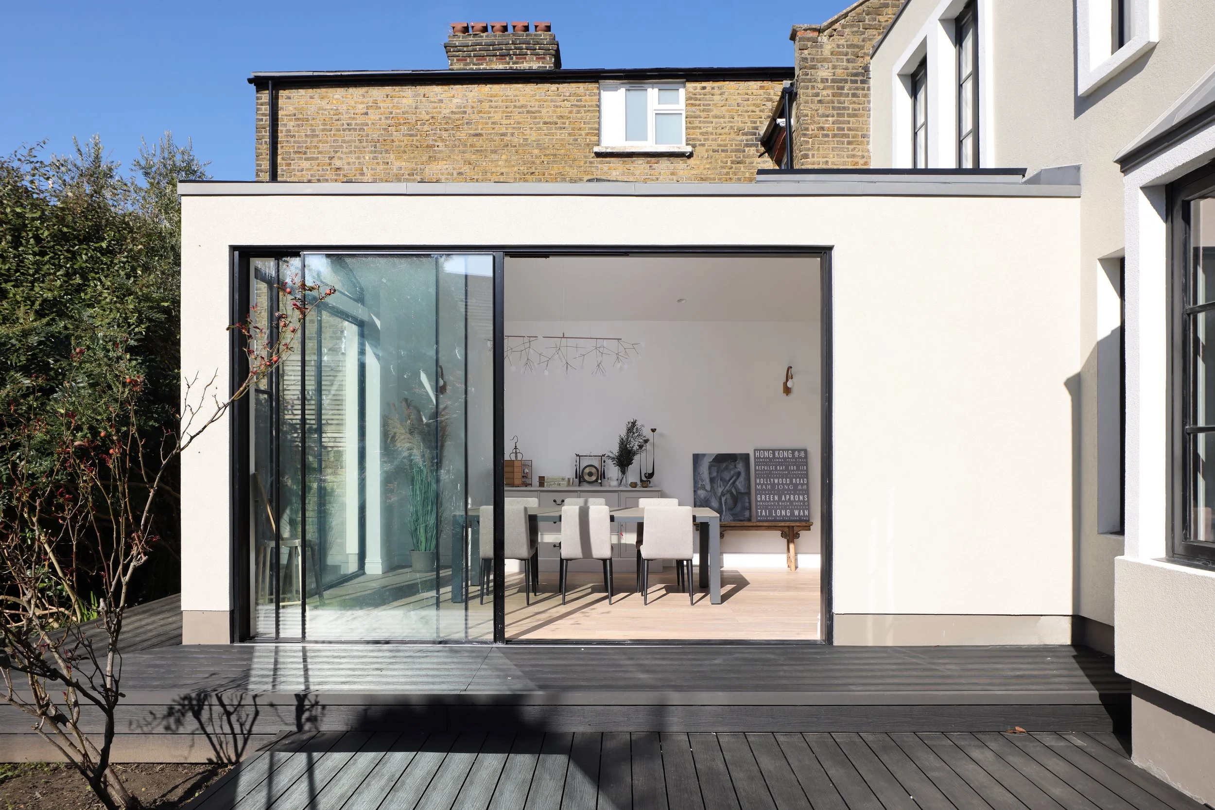 New modern house extension with large glass sliding doors revealing a dining room with six beige chairs around a table, white walls, and contemporary decor.
