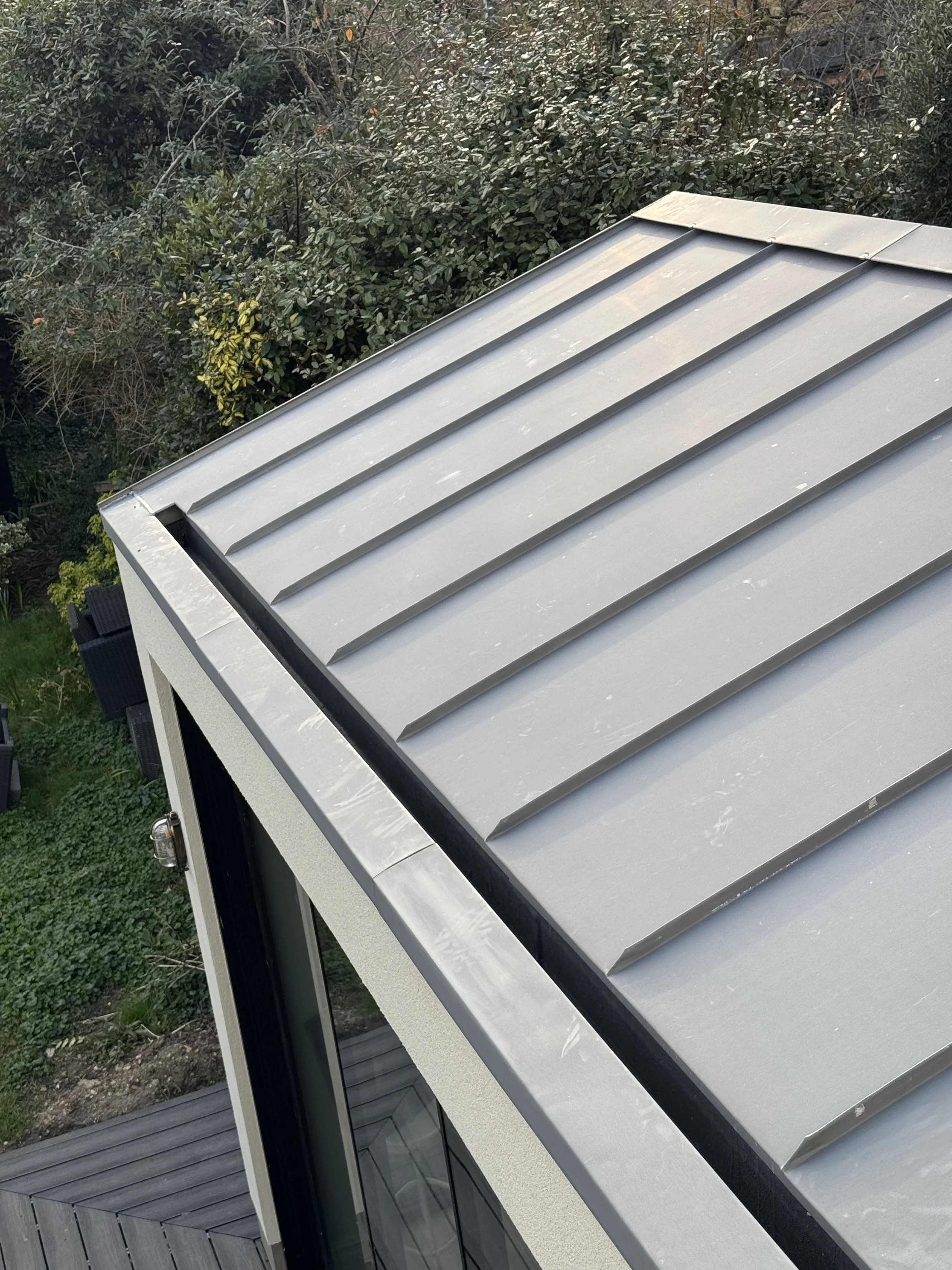 A metal roof with standing seams on a modern building, seen from above, with a background of trees and greenery.