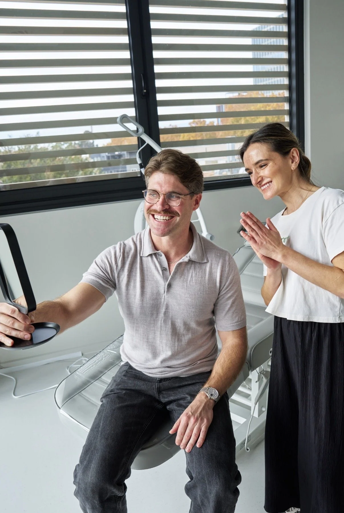 A man with glasses and a woman smiling. The man is sitting on a  examination chair, and the woman is standing next to him, smiling with her hands clasped. The setting appears to be a healthcare or dental clinic with large windows and medical equipment in the background.
