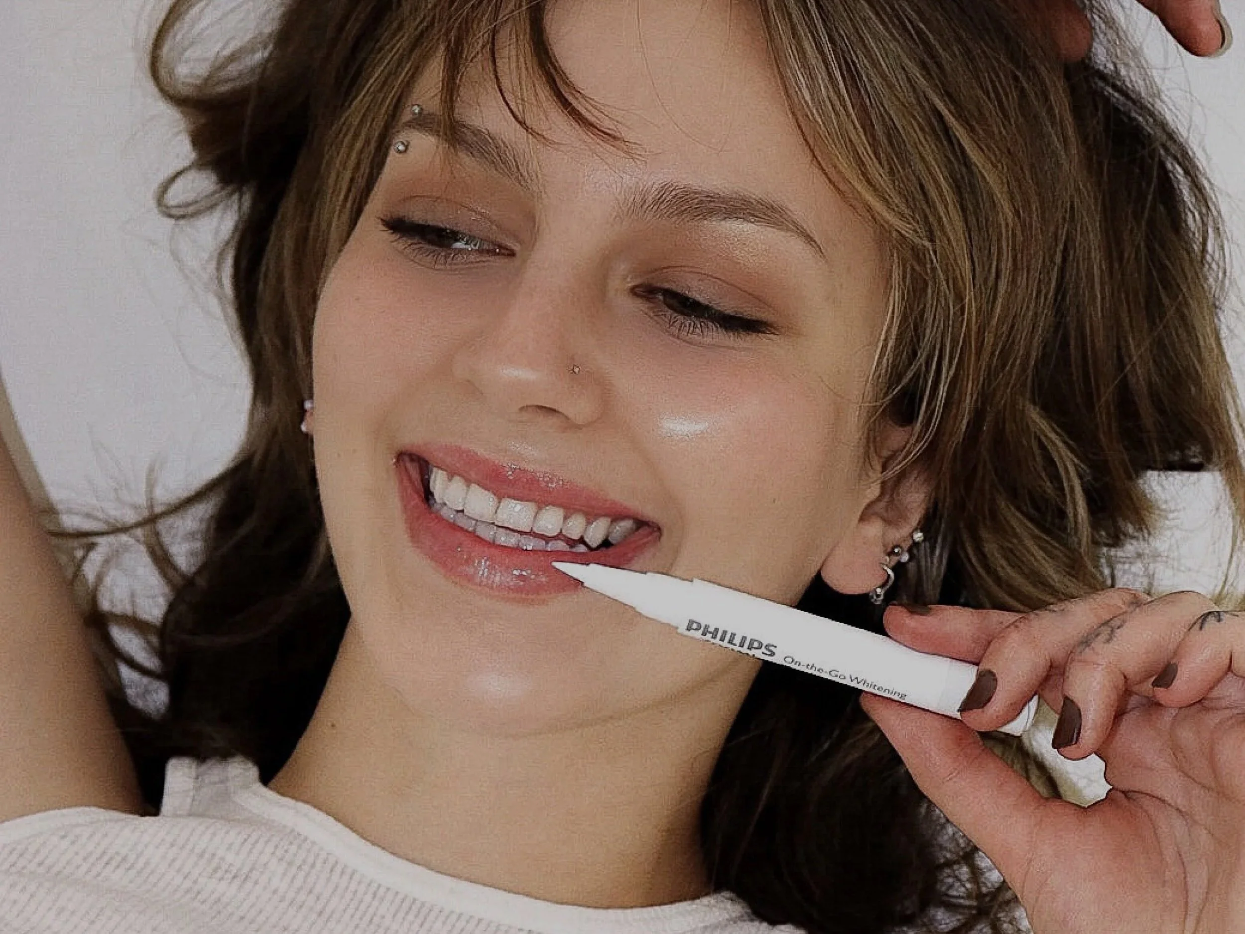 A woman with brown hair and piercings is smiling while holding a whitening pen near her teeth.