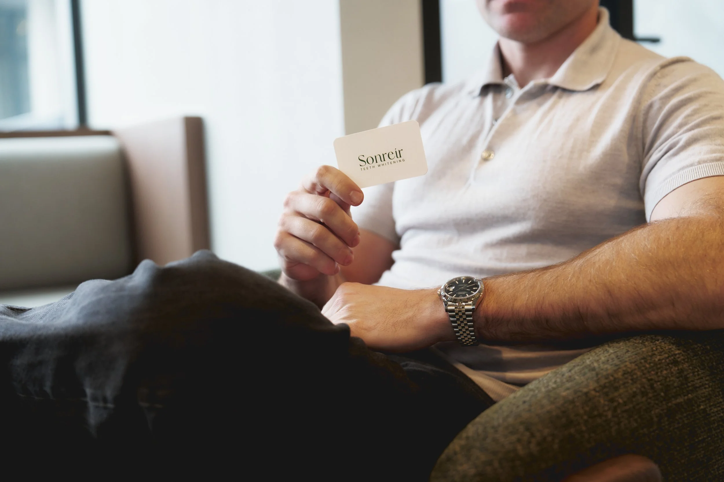 A man wearing a beige polo shirt and a silver wristwatch is holding a business card that has 'Soreir Teeth Whitening' written on it. He is sitting at a table with a laptop in front of him.