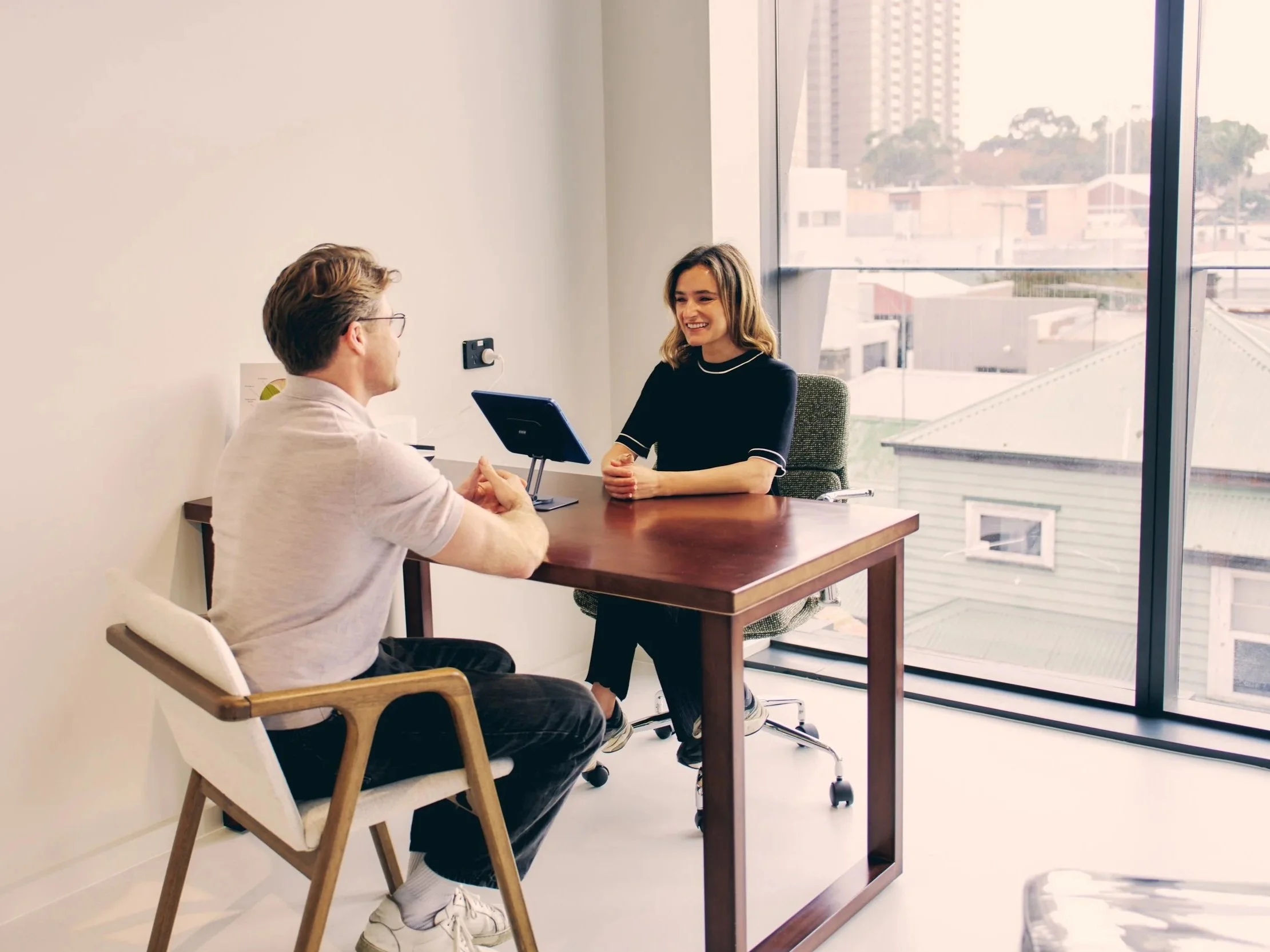 A man and a woman sitting across from each other in a modern office with large windows and city view, engaged in conversation.