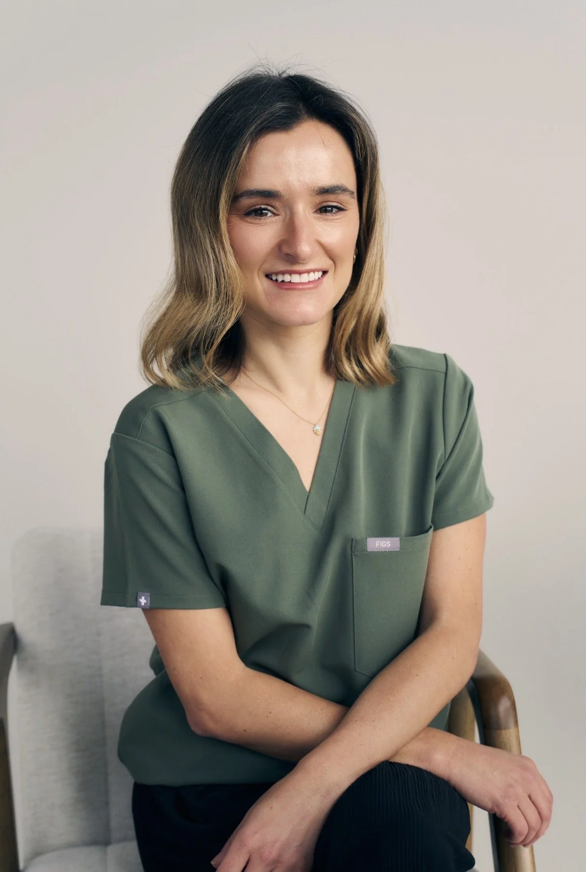 A woman with shoulder-length wavy hair, wearing green medical scrubs, sitting on a beige chair against a plain wall, smiling at the camera.