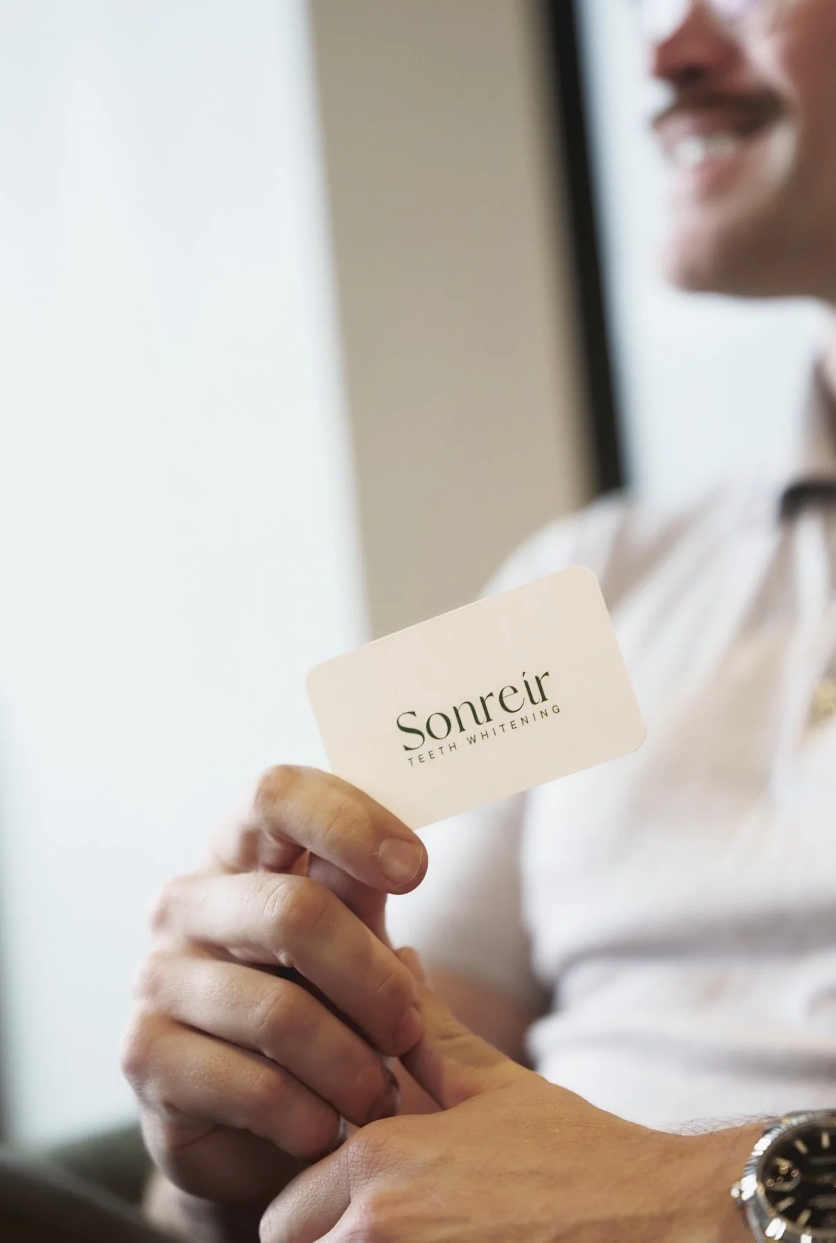 Person holding a business card that says "Sonreír Teeth Whitening" while smiling, with part of their face and hand visible in the background.