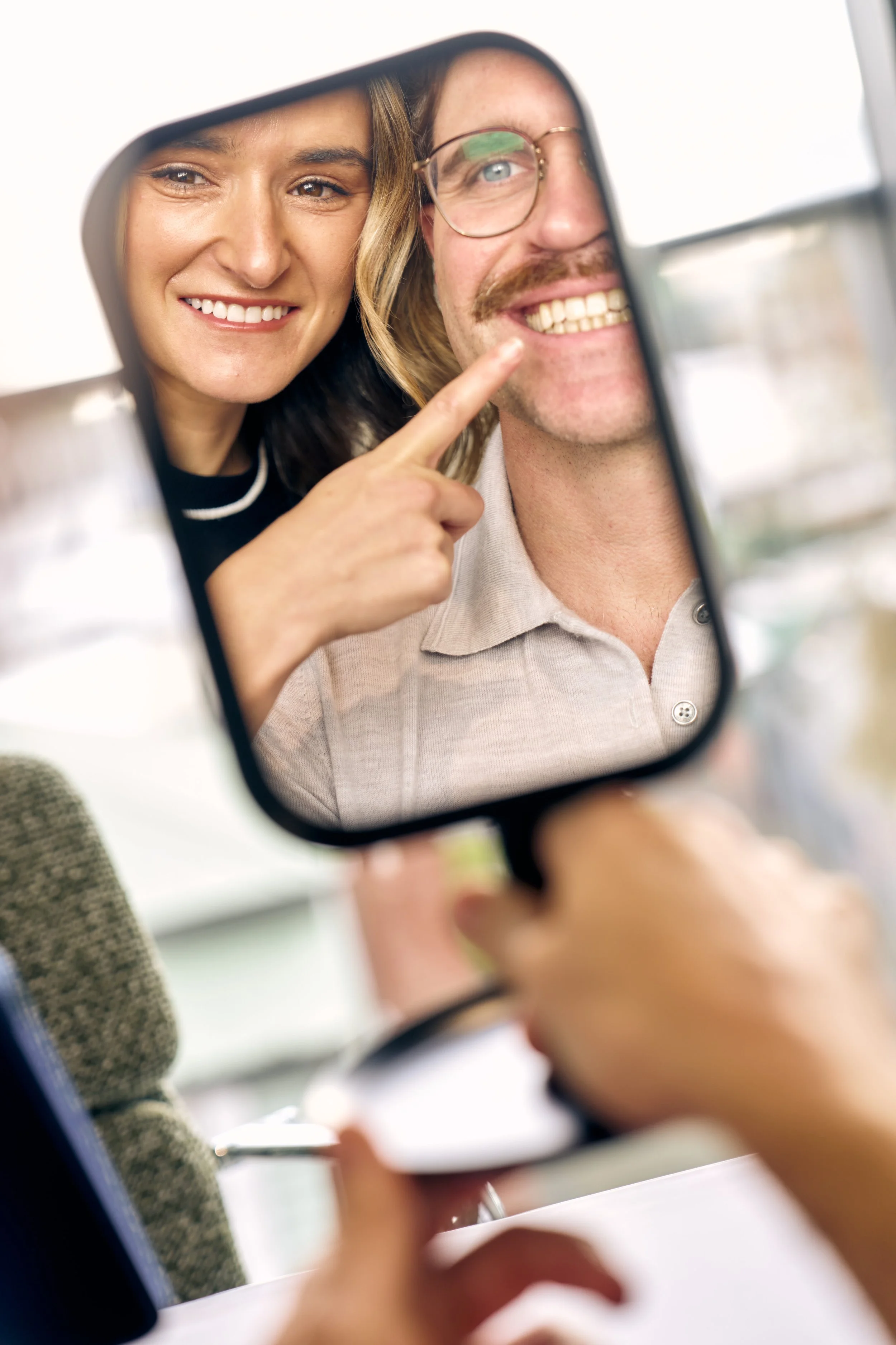 Two people looking into a mirror, smiling, with one pointing at the other's teeth.