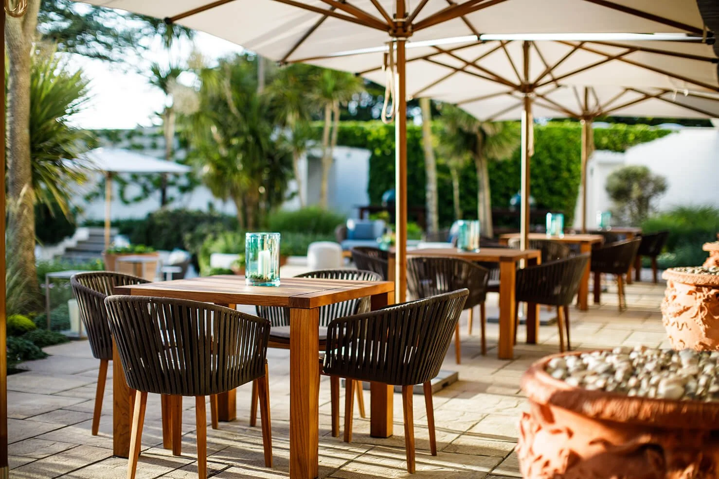 An outdoor patio with wooden tables and black chairs, decorated with glass candle holders, shaded by large beige umbrellas, surrounded by lush greenery and trees.