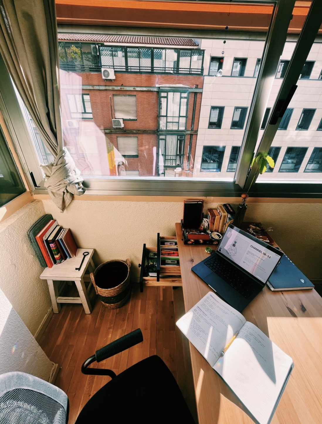 Sunlit home office with open window, desk, open notebook, laptop, books, and decorative items.