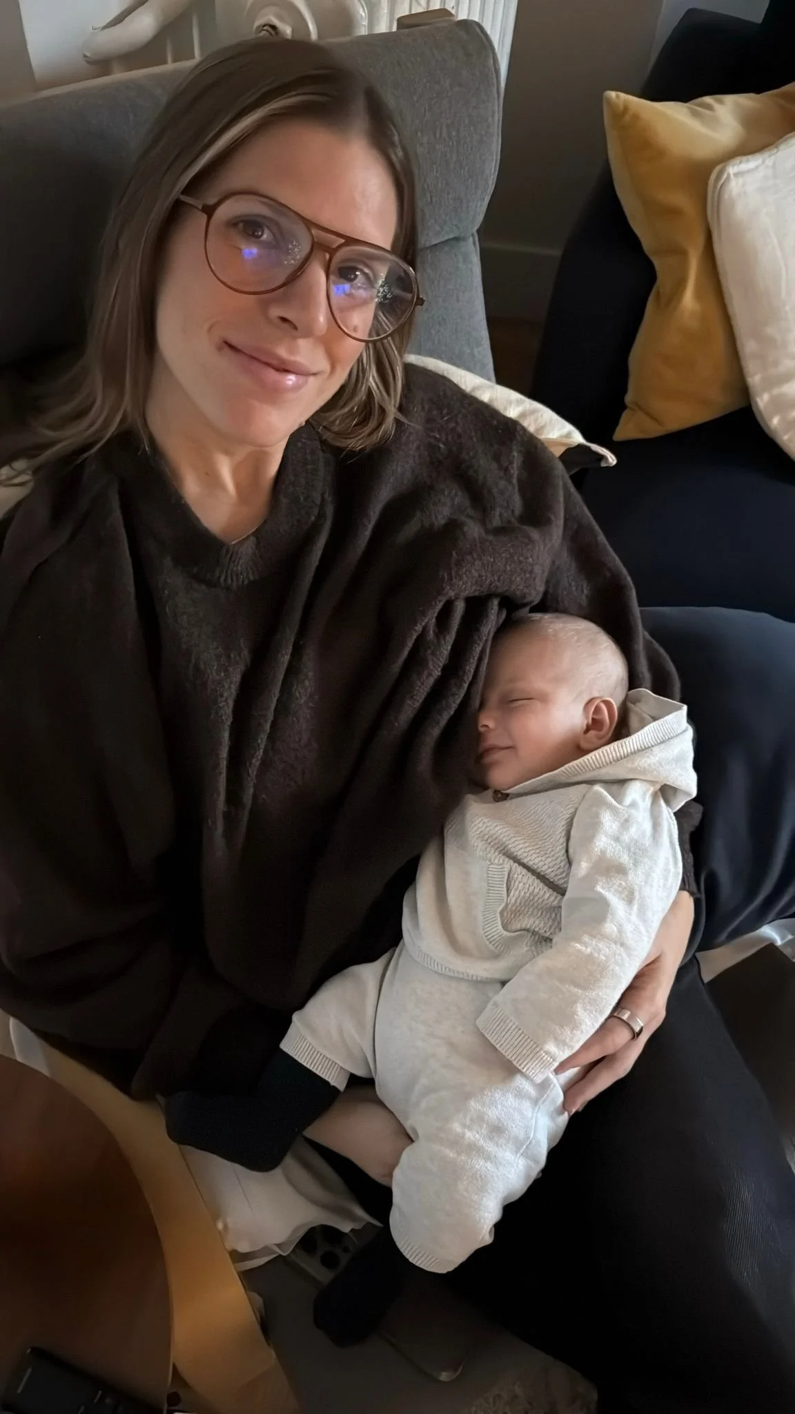 A woman with glasses smiling and a sleeping baby in her arms on a sofa with cushions.