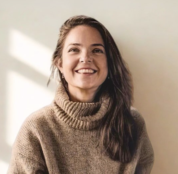 A woman with long brown hair smiling while wearing a beige turtleneck sweater indoors with sunlight coming through a window.