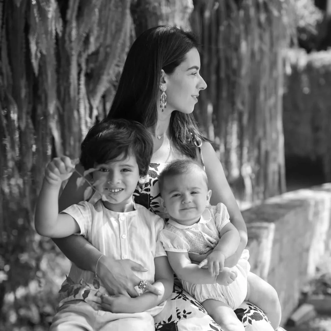 A woman sitting outdoors with two young children on her lap, all smiling and enjoying a sunny day.