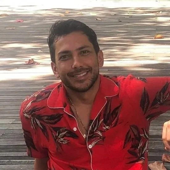 A man smiling, wearing a red Hawaiian shirt with black and brown leaf patterns, sitting on a wooden deck with fallen leaves around.