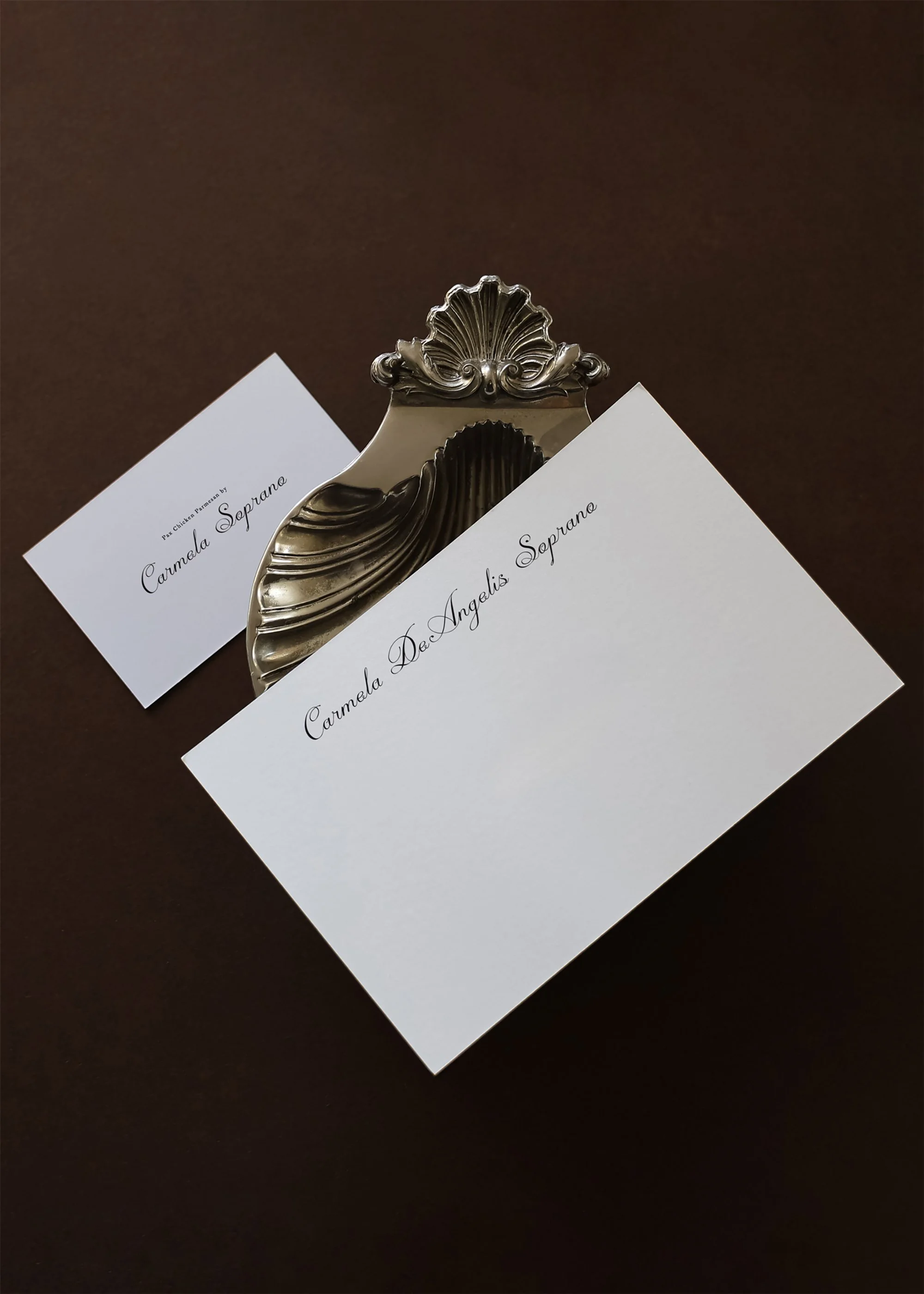 Twee witte kaarten met de namen 'Carmela Dec Ангелis Soprano' en 'Carmela Soprano' liggen naast een zilveren sierlijke naamplaat op een donkere achtergrond.