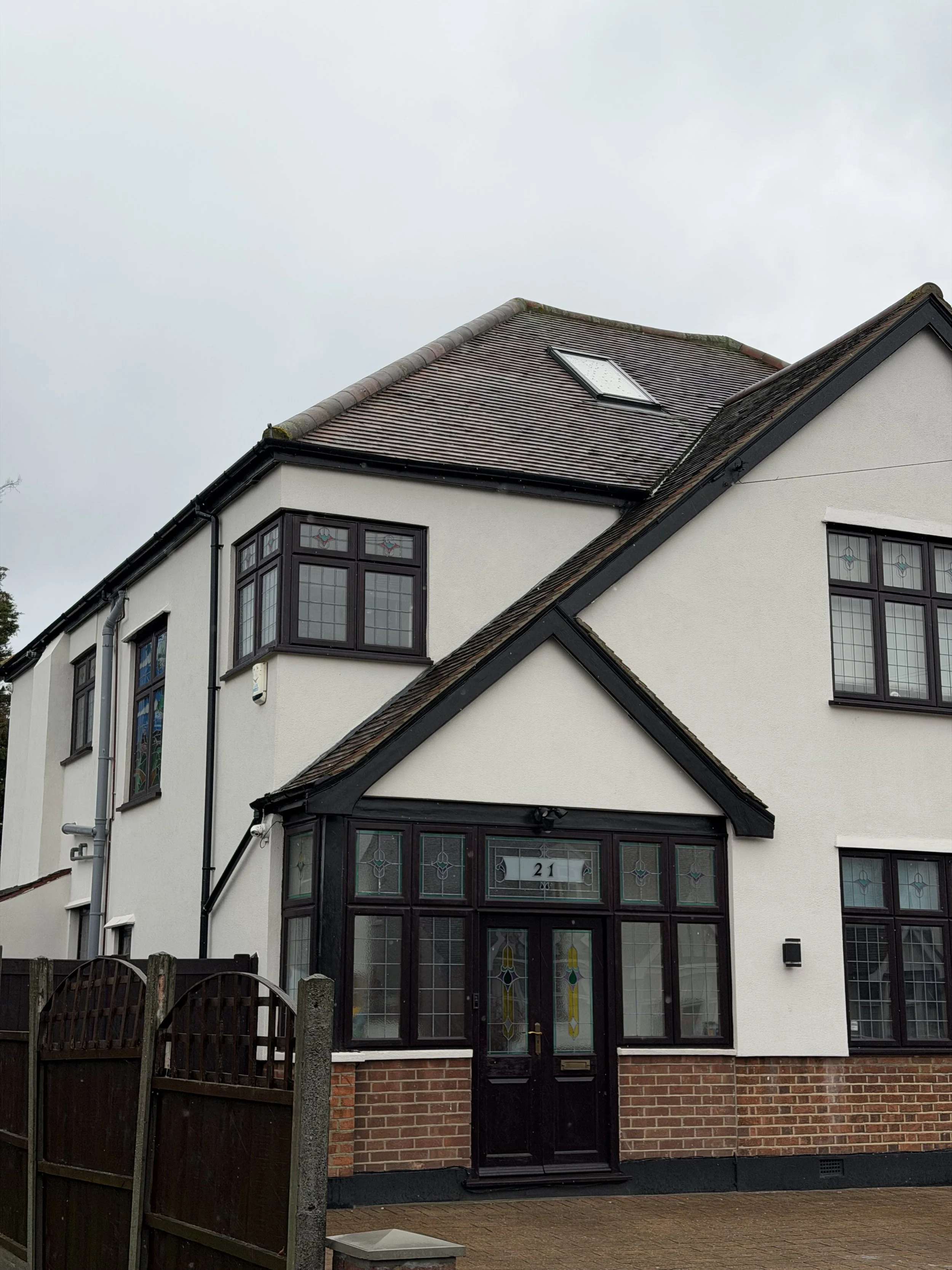 A two-story house with a white exterior, black window frames, and a brick base. The house has a gabled roof with a skylight. The front porch features stained glass windows and a black door with house number 21. A wooden fence is in front of the house under an overcast sky.