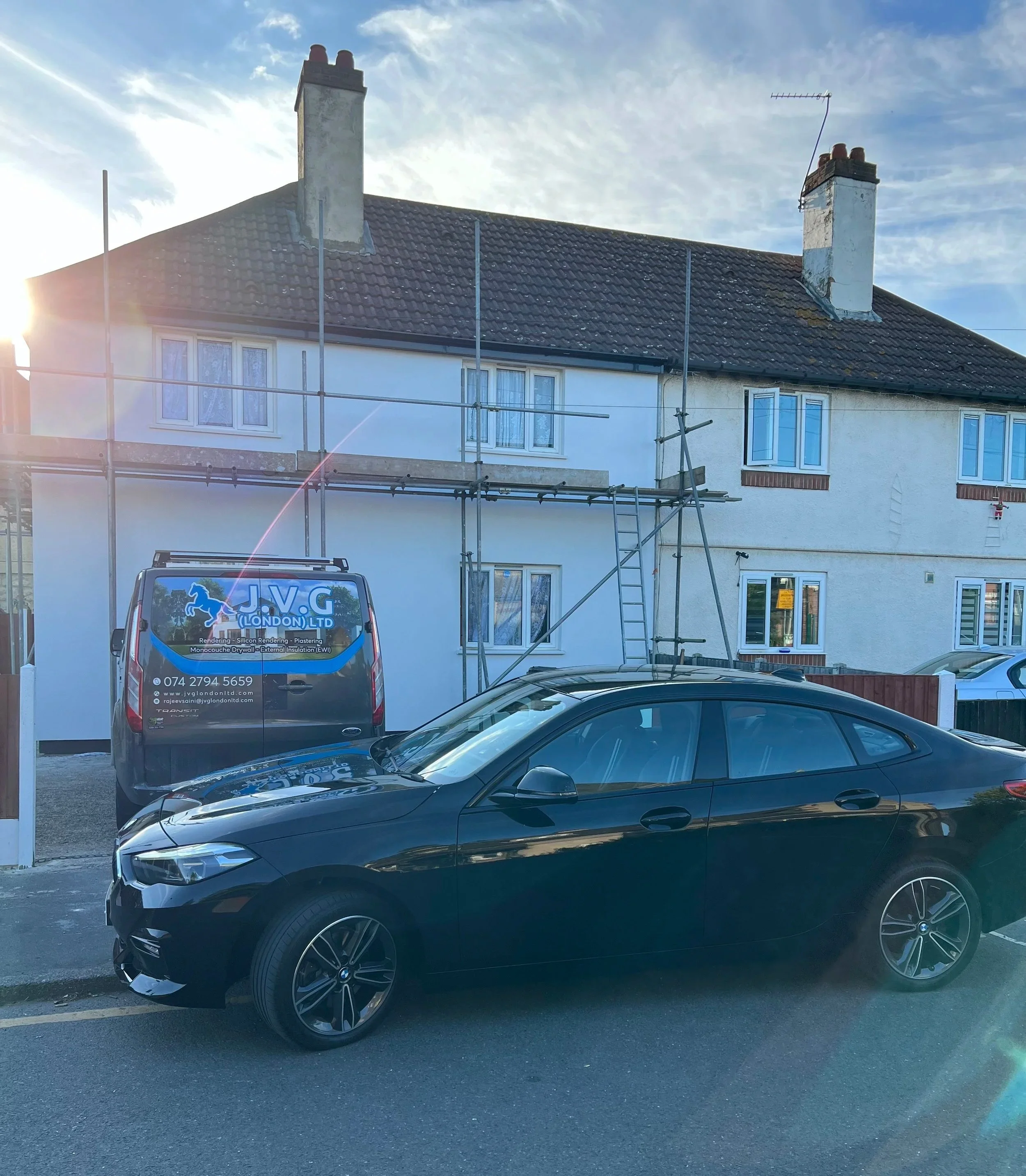 A black car parked on the street in front of a house under renovation, with scaffolding and a van belonging to a construction company nearby. The house has white walls, a tiled roof, and two chimneys.