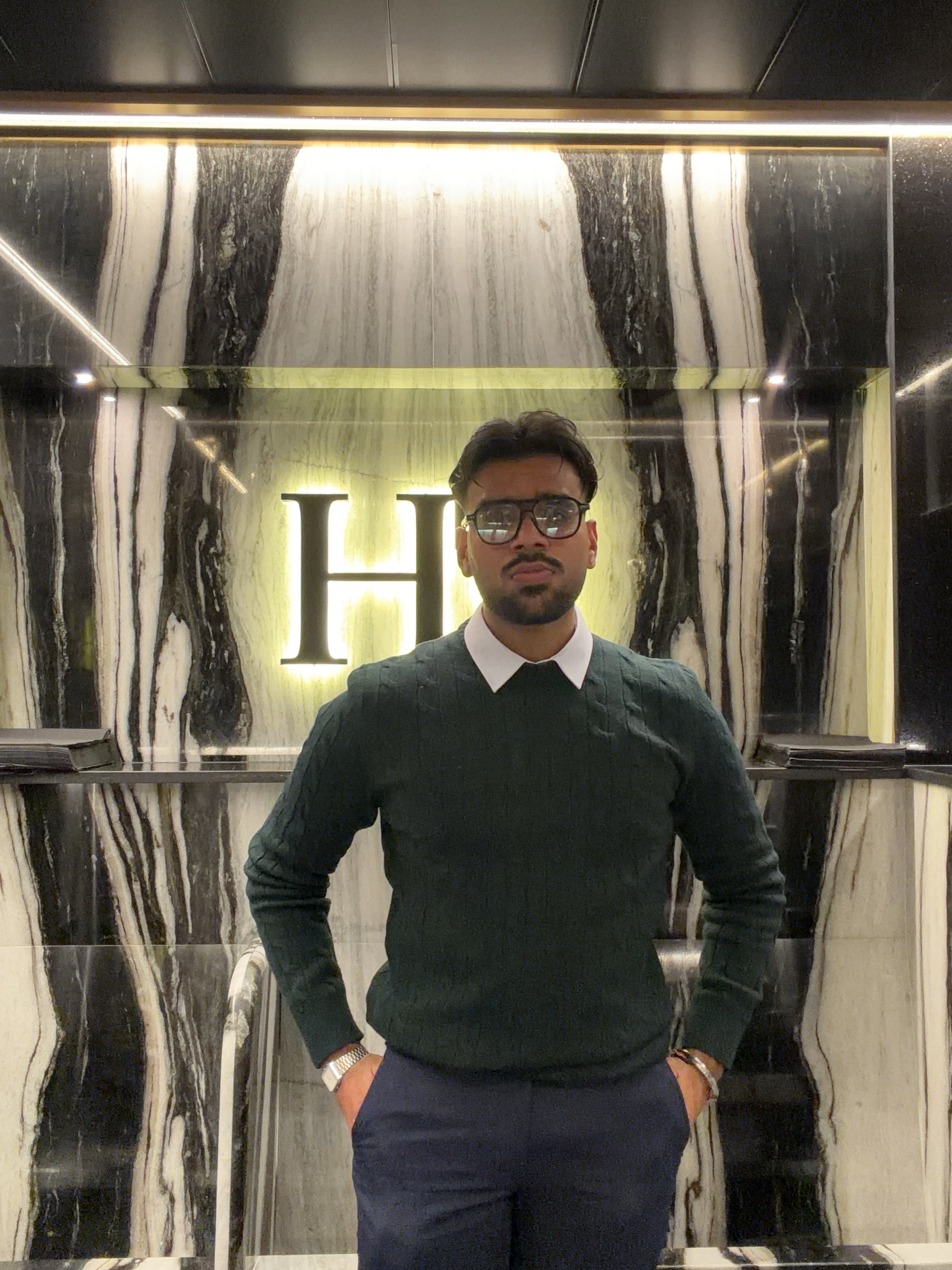 A man wearing glasses, a dark green sweater, and navy pants standing in front of a black and white marble wall with illuminated letter 'H' behind him.