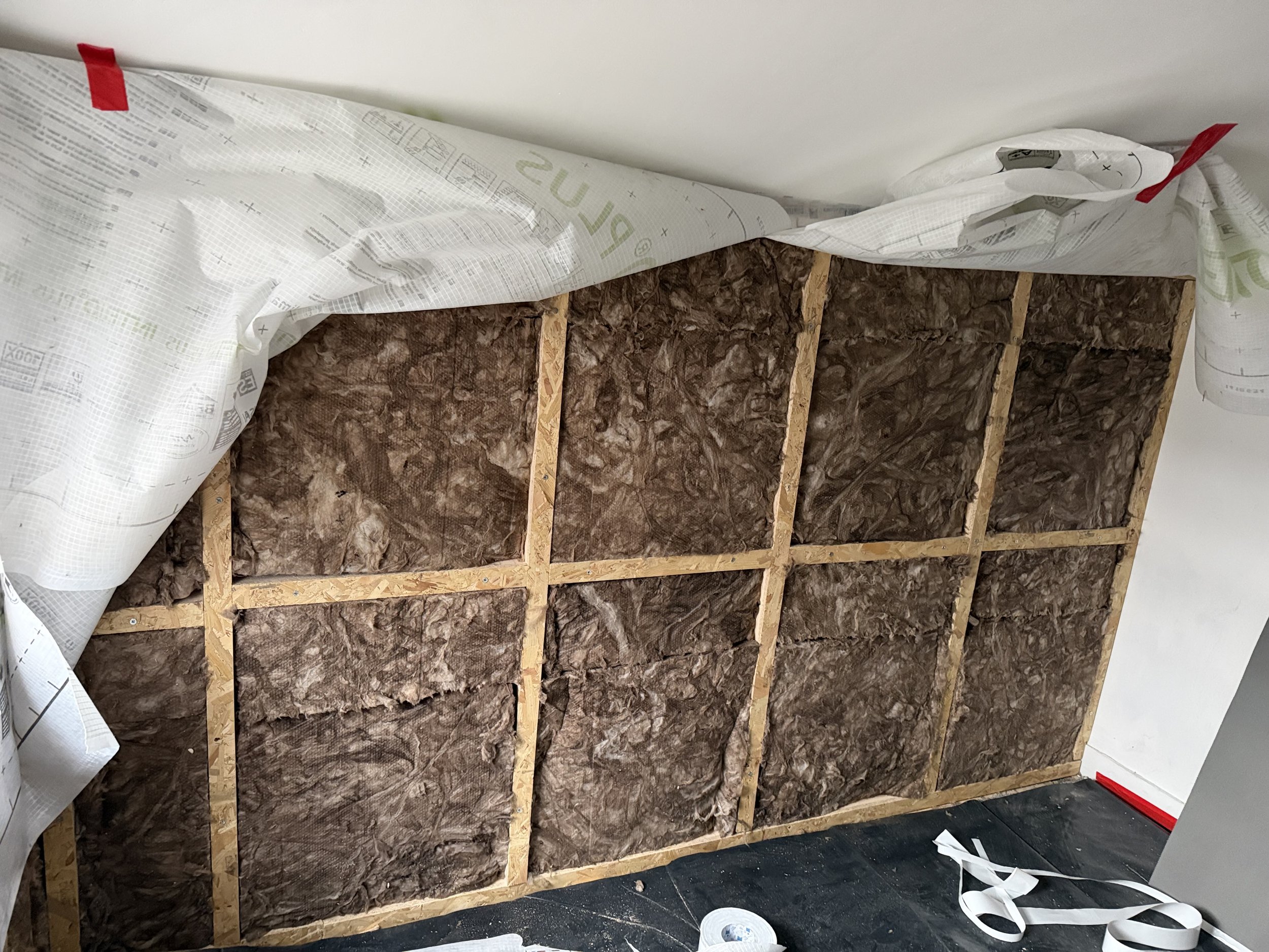 In-progress insulation installation showing brown fiberglass insulation within wooden framing on a wall, with some plastic vapor barrier partially peeled back.