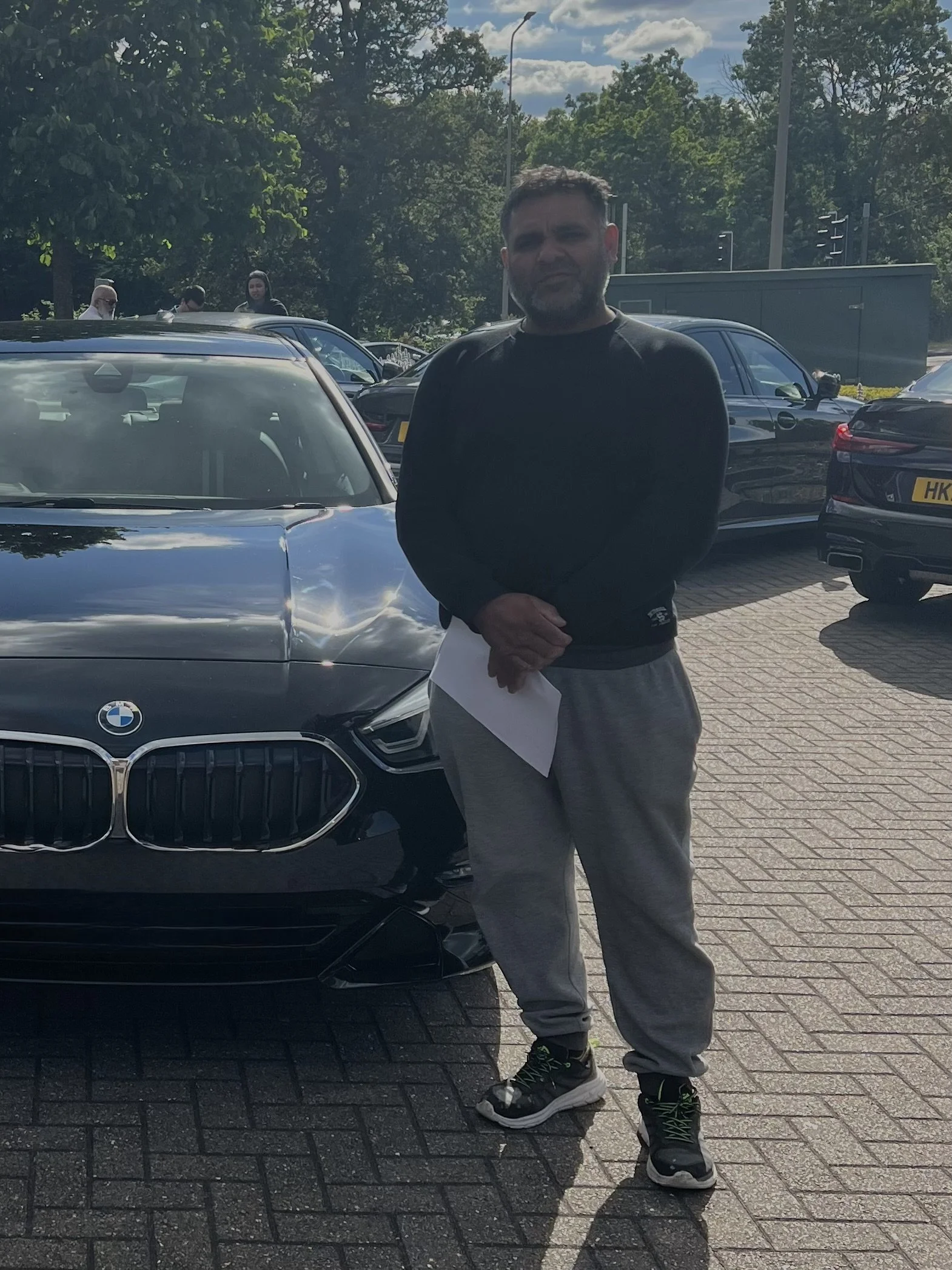 A man standing in front of a black BMW car, holding a piece of paper, with a parking lot and other cars in the background, under a partly cloudy sky.