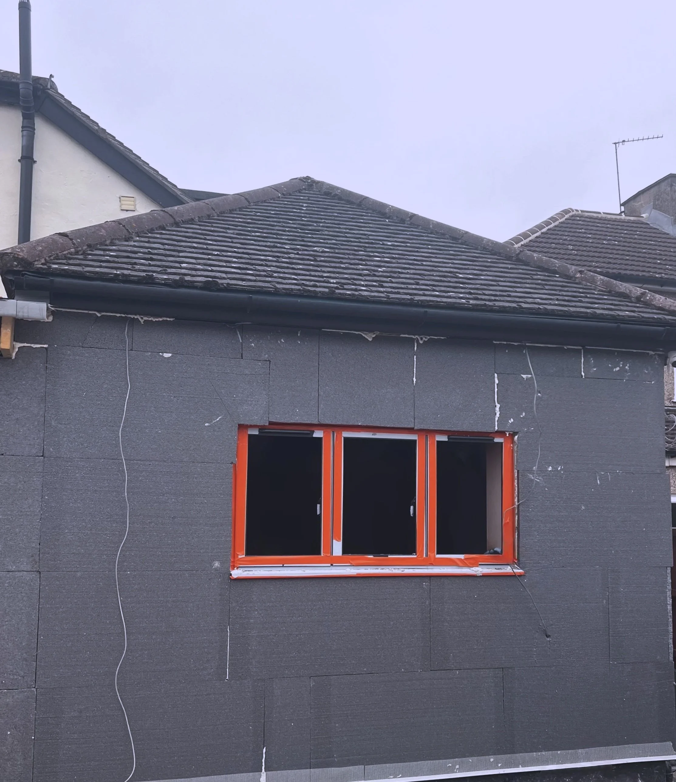 Construction site of a building with gray exterior wall, a window with orange frame, and a shingles roof.