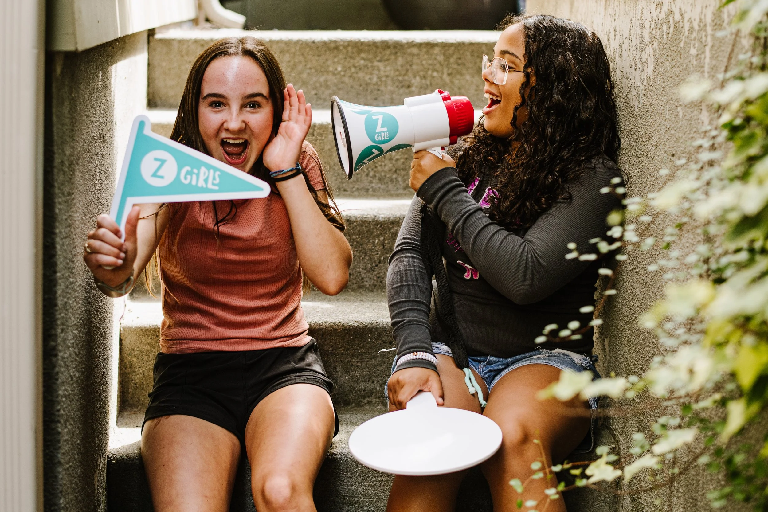 A girl in a grey sweatshirt pretends to scream into a megaphone. The girl to the left of her pretends to scream in reaction to the noise.