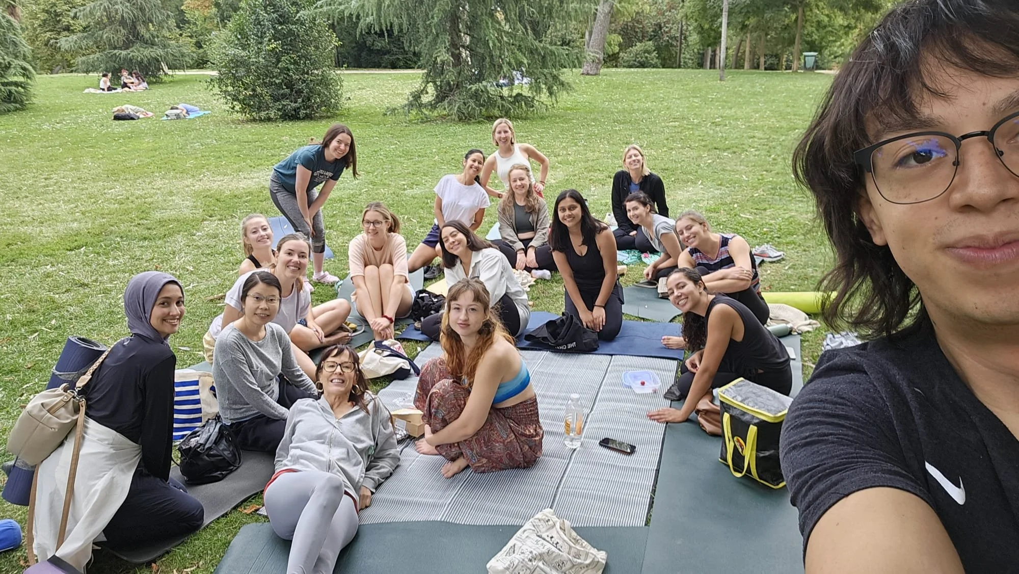 A group of people, primarily women, gathered on mats in a park, taking a group selfie with some people sitting, kneeling, or crouching on the grass, and others standing behind, all smiling and looking at the camera, with trees and more groups in the background.