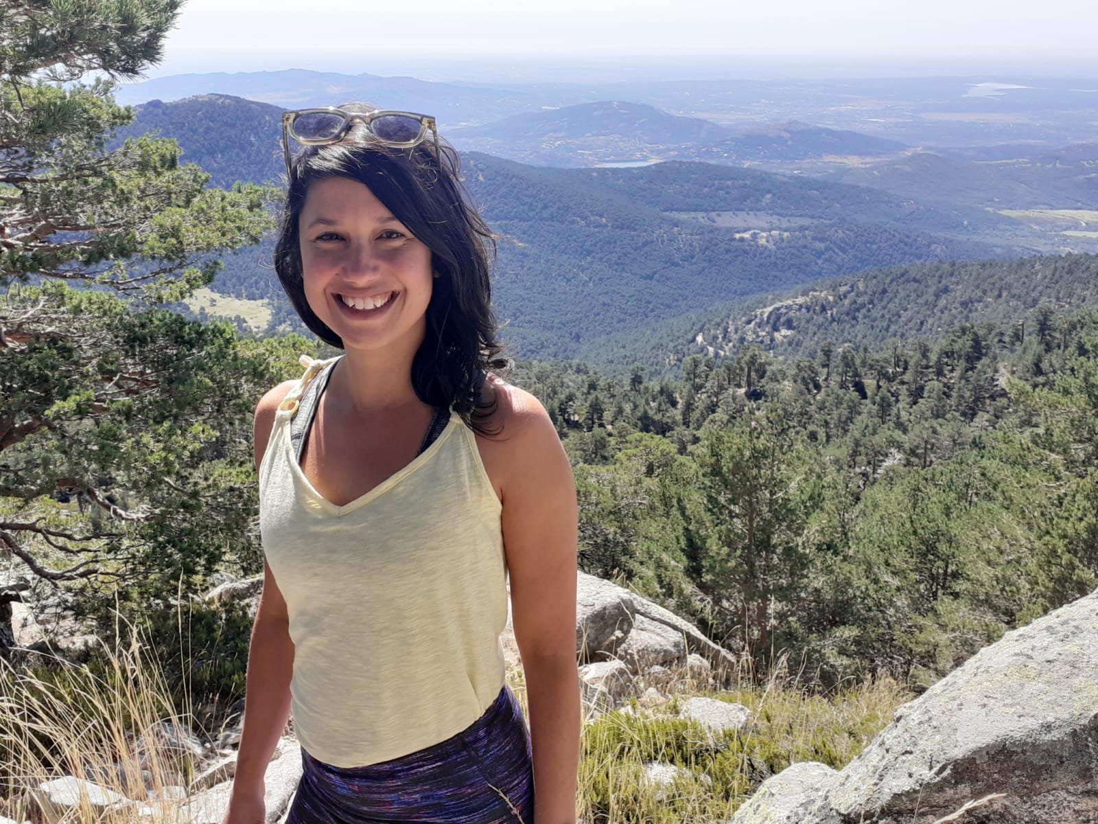 Person standing outdoors on a mountain with forested landscape and distant mountains in the background, smiling and wearing a light-colored tank top and sunglasses on their head.