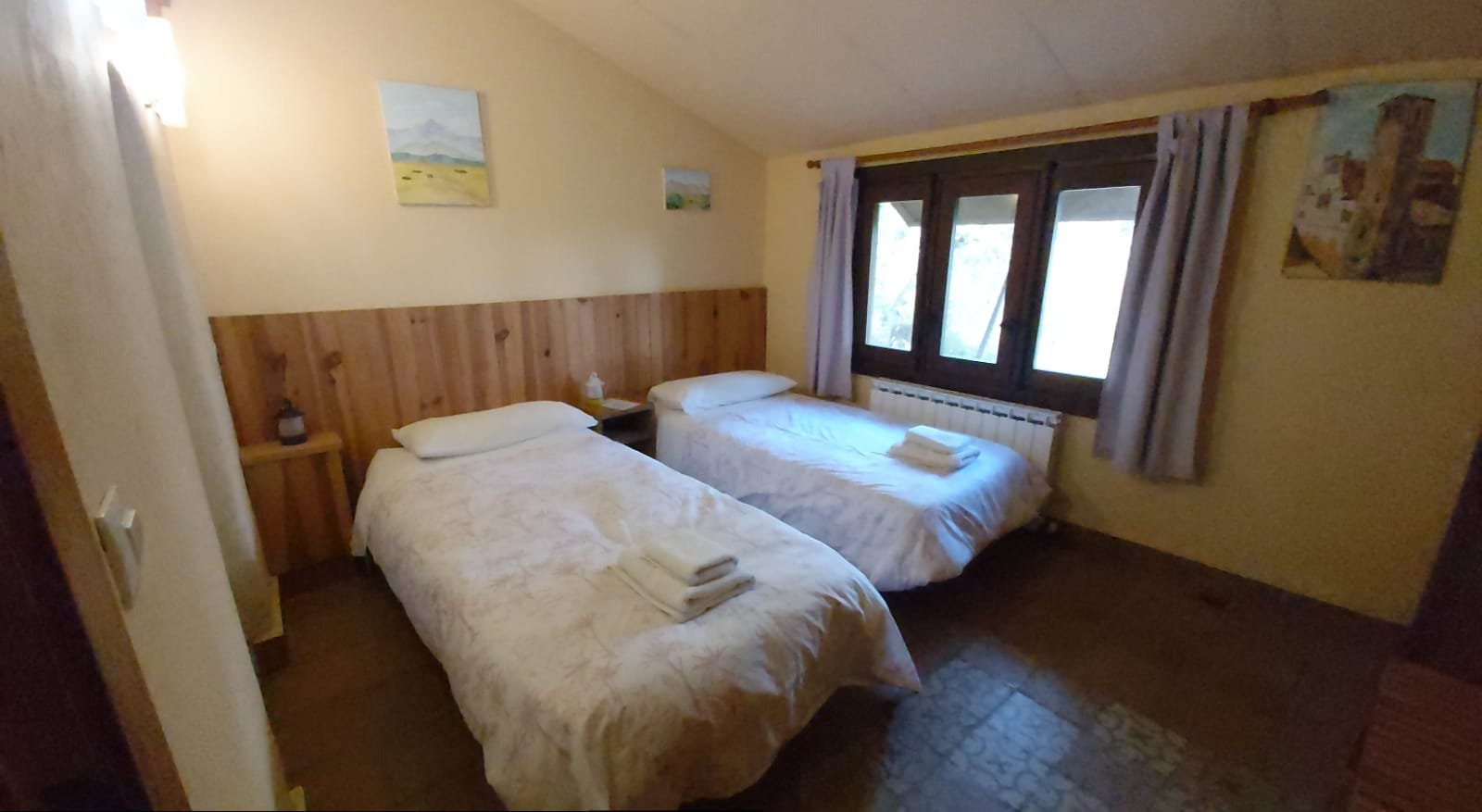 A cozy bedroom with two single beds, each with white bedspreads and pillows, arranged against a wooden paneled wall. The room has a window with curtains, a small bedside table with a lamp, and wall paintings for decoration.