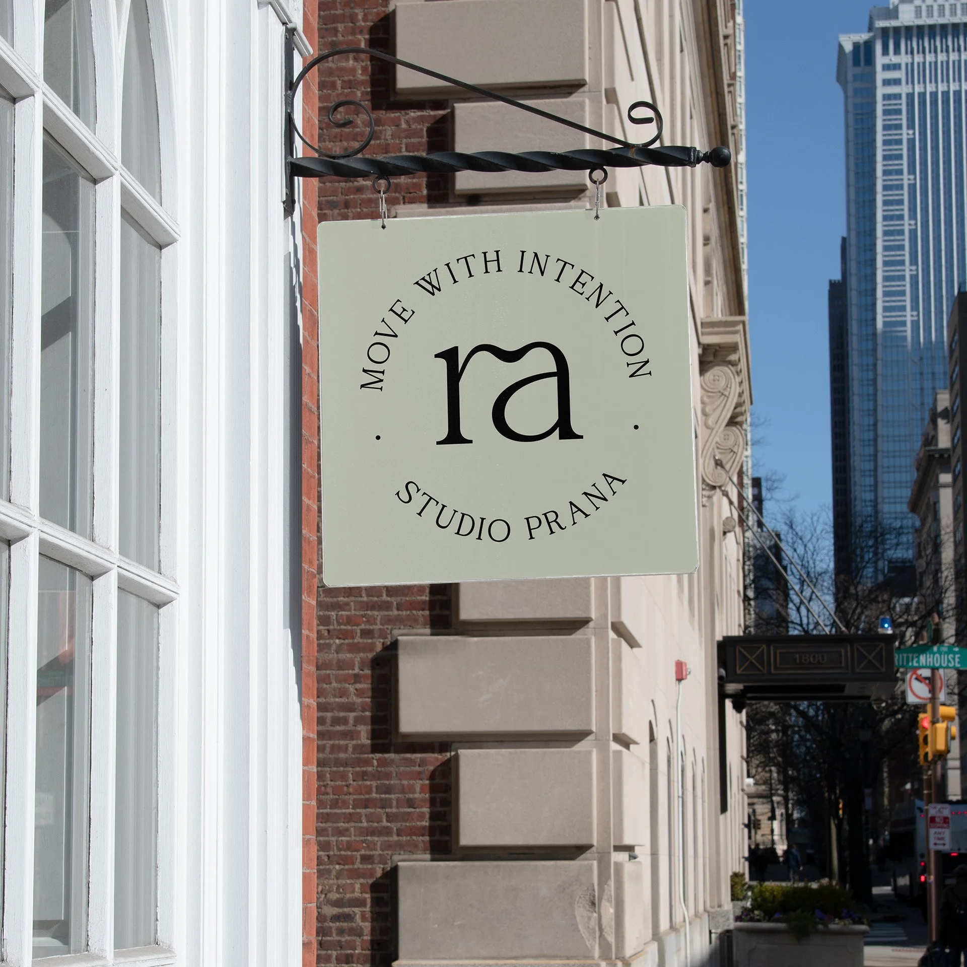 Street sign reading 'Move with Intention Studio Prana' with a large 'ra' in the center, hanging on a building in an urban area with skyscrapers in the background.