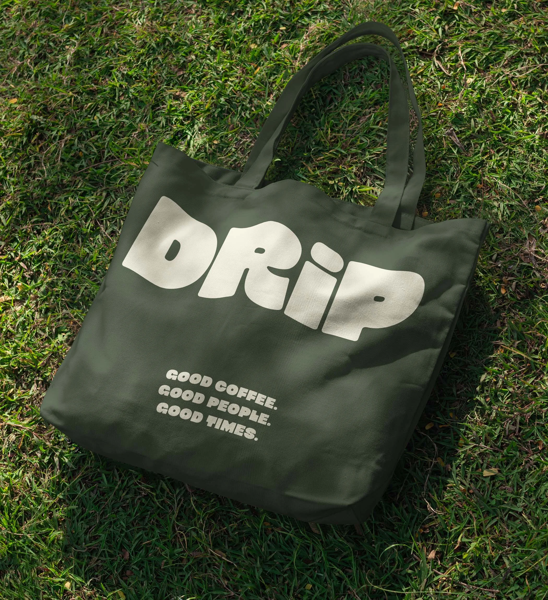 Green tote bag with white text that says 'DRIP' and smaller text that reads 'Good Coffee, Good People, Good Times,' lying on grass.