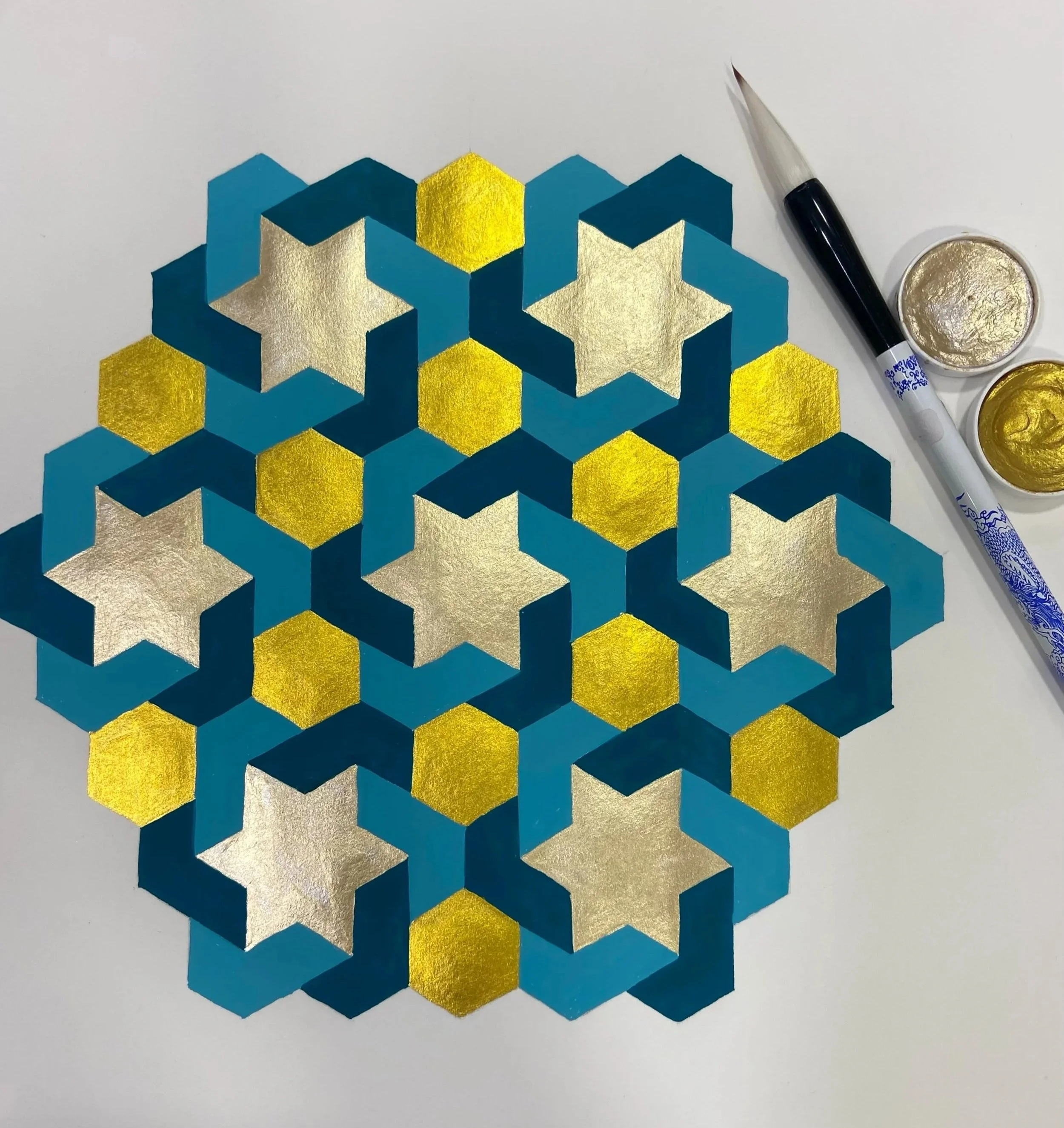 Close-up of a geometric artwork with 3D hexagonal shapes in blue, gold, and white, with gold and silver metallic finishes. A black and white pen and small containers of gold and silver metallic paint are on the right side of the artwork.