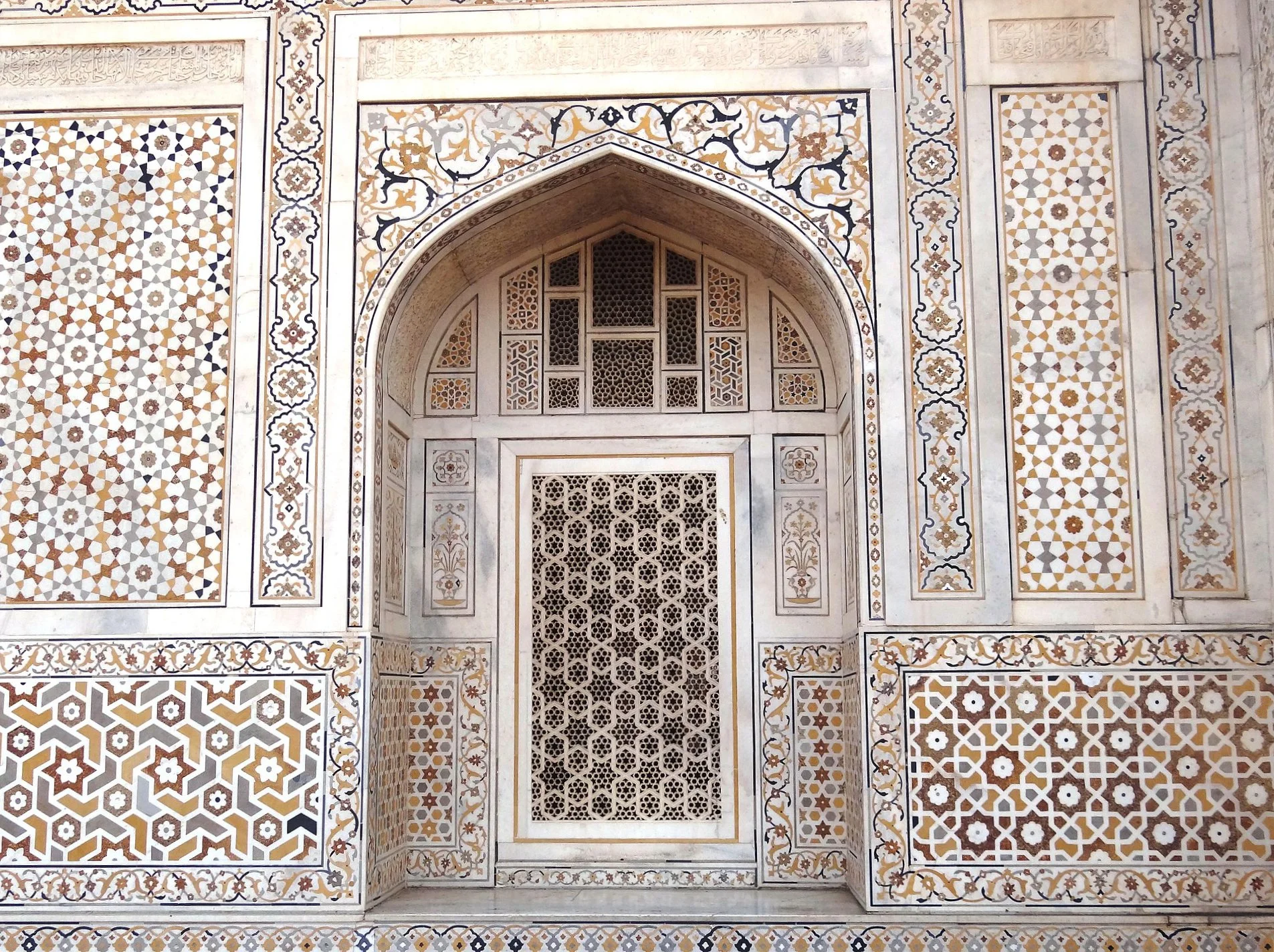 Close-up of ornate white marble wall with intricate geometric and floral inlay patterns in gold, black, and gray, featuring a decorative arch and window with lattice design.